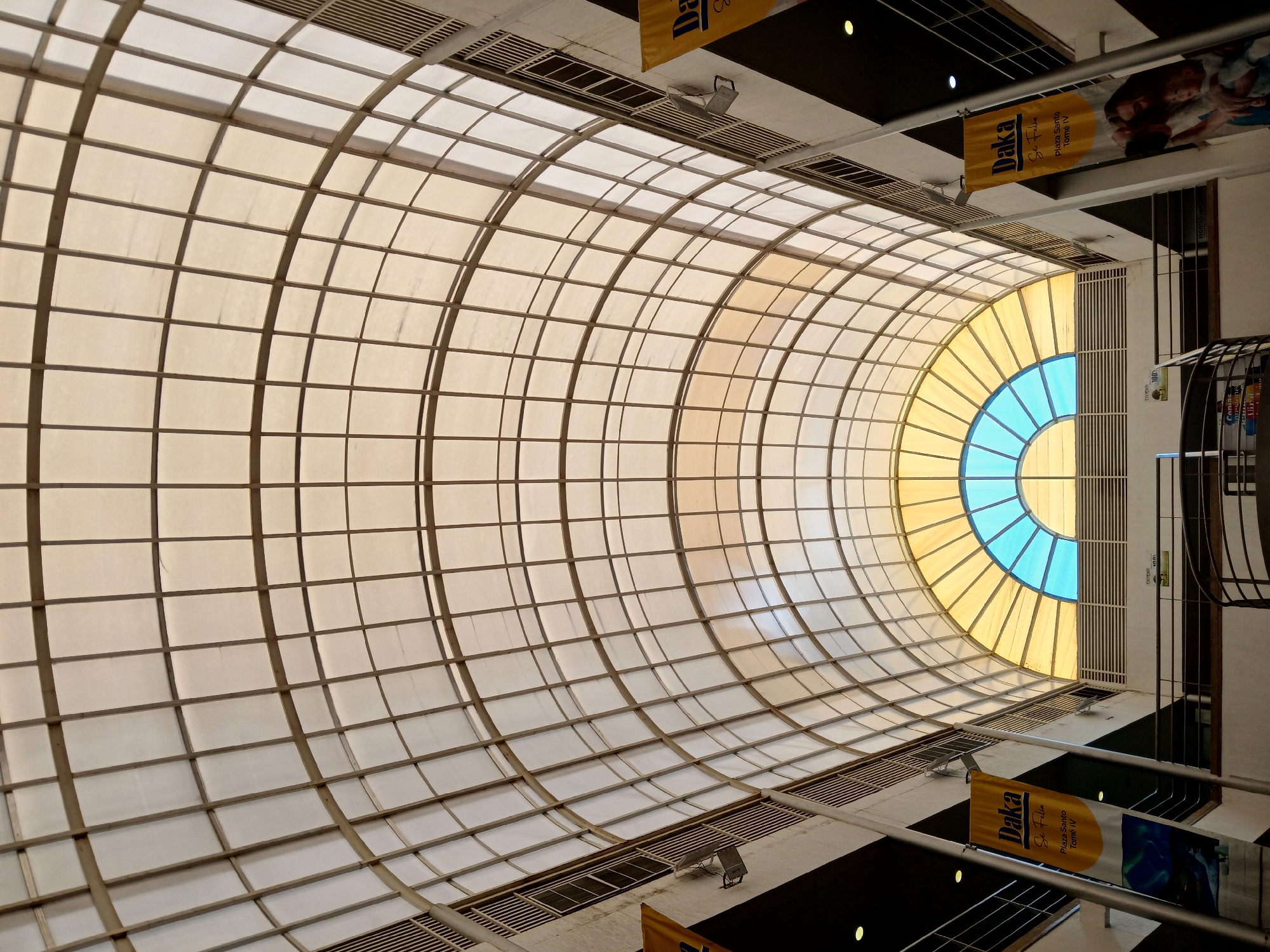 Here's a detailed description of the image:
**Setting:**
The image is taken from below, looking upwards into a large, curved skylight. The skylight is constructed with a grid of metal supports, forming rectangular or square panels that are filled with translucent material, allowing light to filter through.
**Architecture:**
The skylight structure is a prominent architectural feature, creating a tunnel-like effect. It appears to be a significant design element within a commercial building or shopping mall. At the end of the structure, a semi-circular opening allows a glimpse of the sky, which appears blue.
**Details:**
* The interior of the building is visible around the skylight. There are walls and possibly balconies or levels.
* Yellow banners, with the name "Daka Se Feliz Plaza Santo Tomé IV" and images are hanging from the ceiling or upper levels, indicating a business promotion or advertising inside the mall
* There are also ventilation grates and lighting fixtures visible near the edges of the skylight structure.
**Location:**
The banner says "Plaza Santo Tomé IV" which is located in Ciudad Guayana, Venezuela.
