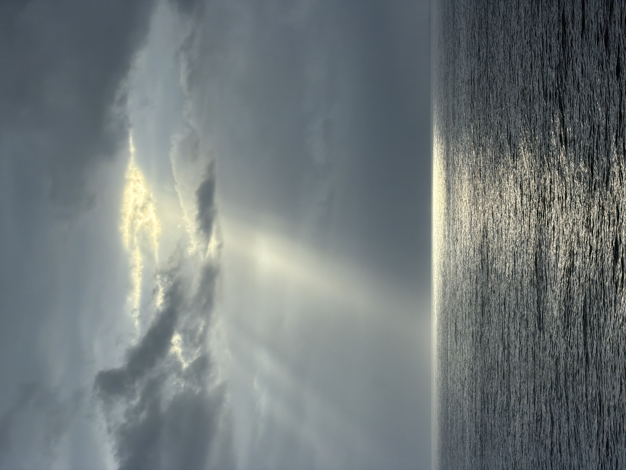 The image displays a dramatic seascape or large body of water beneath a cloudy sky, presented vertically but interpretable horizontally as a landscape. The sky is dominated by heavy, dark grey clouds, but a significant break in the cloud cover allows brilliant sunlight to stream through. These crepuscular rays, appearing golden and white, illuminate portions of the clouds and extend downwards.
The light strongly reflects on the surface of the water, creating a wide, shimmering path of intense brightness that stretches from the horizon towards the viewer. The illuminated water shows a textured surface with many small, reflective ripples or waves. The rest of the water body appears darker and calmer. A clear, straight horizon line divides the expansive sky from the water.
The scene suggests either a sunrise or sunset, or a moment when the sun breaks through after an overcast period, due to the dramatic light and cloud interaction. The weather is partially cloudy with direct sunlight. No specific geographical landmarks are visible, indicating an open natural environment. No text is visible in the media file.