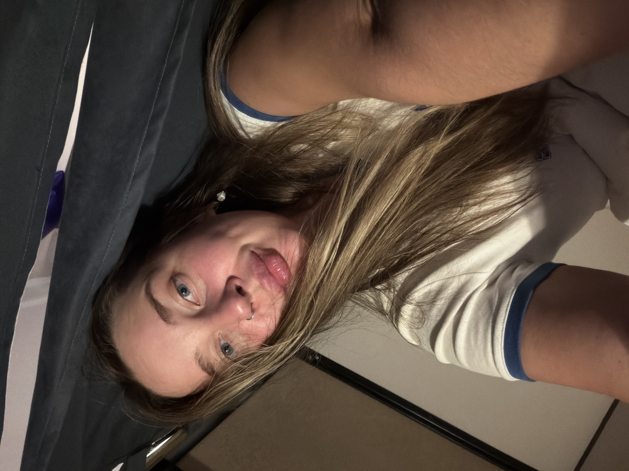 A selfie of a woman with fair skin, blue eyes, and blonde hair. She is wearing a white t-shirt with blue trim. The background is dark and includes a curtain. The image does not contain information to identify the location as Moscow, Russia.