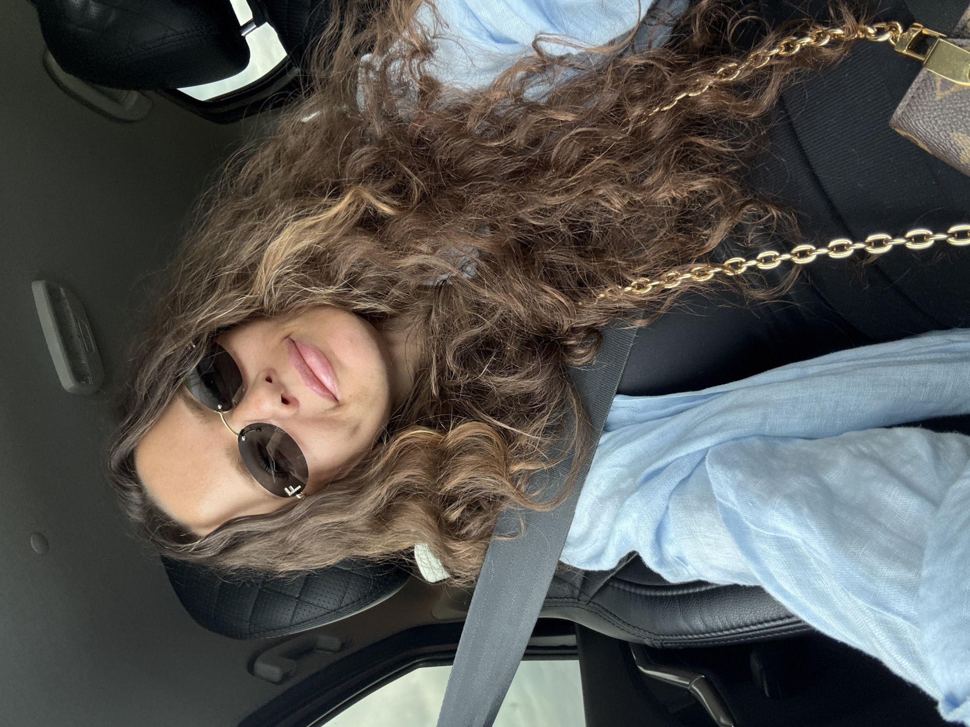 A woman is sitting in the driver's seat of a car. She is wearing sunglasses, a light blue shirt, and a black top. A gold chain purse is visible on her right side. The car interior is visible, including the black leather seats and a gray seatbelt. The background is blurred, suggesting the car is in motion. There is no information about the location in the city of Mokina, Russia.