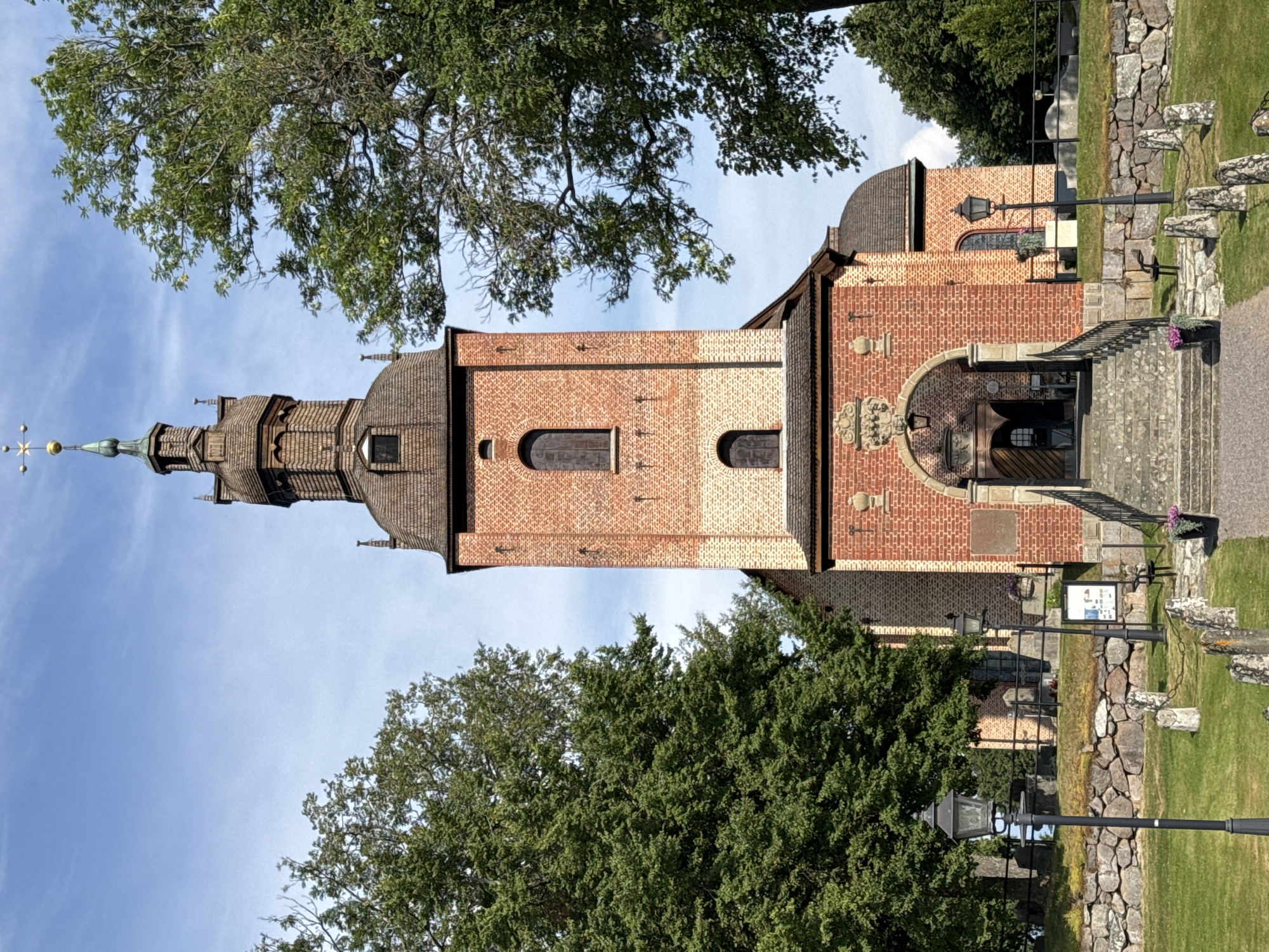 The image shows the church of Sund in Askersund, Sweden. The church is a red brick building with a tall, wooden-shingled tower topped with a spire. The entrance to the church is through a stone archway, with stone steps leading up to the doors. The church is surrounded by a graveyard with gravestones and a stone wall. There are trees on either side of the church, and the sky is blue with some clouds. Two black street lamps are visible in the foreground. The image is taken from a low angle, looking up at the church.