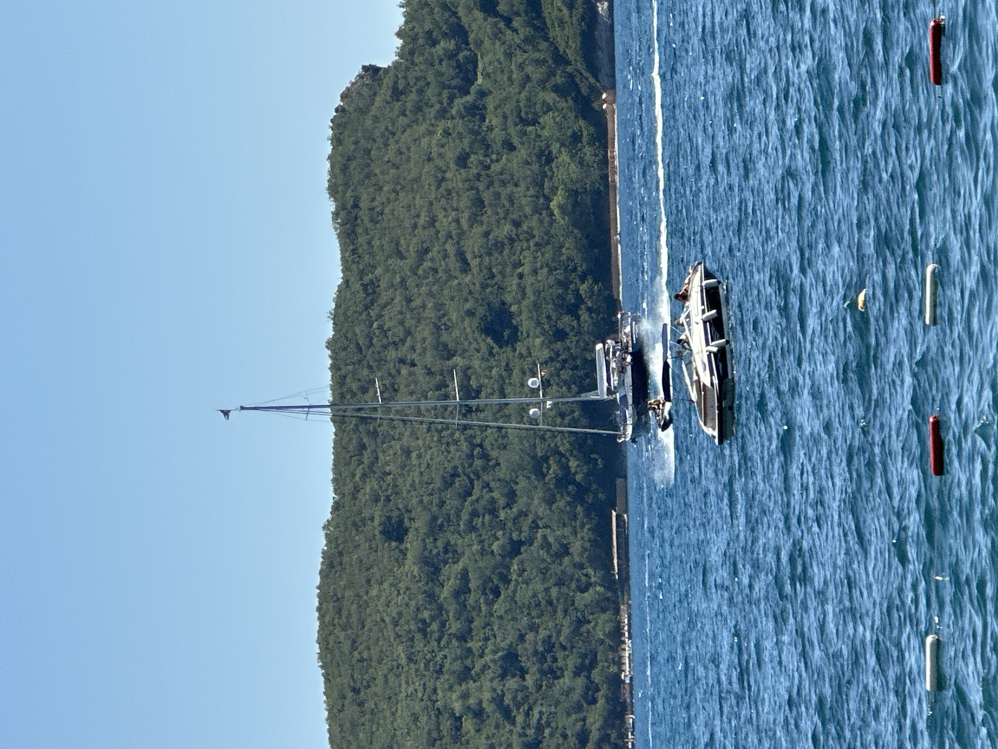 This is an eye-level, medium-shot, vertical photograph captured on a clear, sunny day, likely in Budva, Montenegro. The image depicts a wide expanse of blue, choppy water in the foreground, with a lush, green, tree-covered hill forming the backdrop. Floating buoys with red and white markings are visible on the water's surface. In the middle ground, two boats are in motion. A smaller, white speedboat with at least three people aboard is closer to the viewer and appears to be moving away. Behind it, a larger sailboat with a prominent mast and a black flag at its peak is also moving, creating a wake. In the far distance, a shoreline is lined with structures, possibly hotels or resorts, and more vegetation. The sky is a clear, bright blue, indicating fair weather.