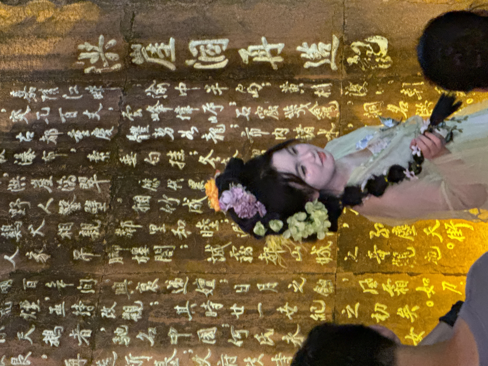 This image shows a group of people in front of a large stone wall covered in carved Chinese calligraphy. The calligraphy is highlighted with a golden paint. In the foreground, a young woman with elaborate hair ornaments, including flowers and braided hair, is dressed in traditional Chinese attire. She is looking to her right, with a slight smile. To the left and right of the woman, parts of other people are visible, also facing the wall. The wall appears to be outdoors, with rough, textured stone. The lighting suggests it might be evening or a dimly lit indoor space. The calligraphy is ancient and weathered, indicating a historical or cultural site.
The specific location is the Yuzhong District of Chongqing, China, known for its many historical sites and stone carvings, including those found on the ancient city walls.