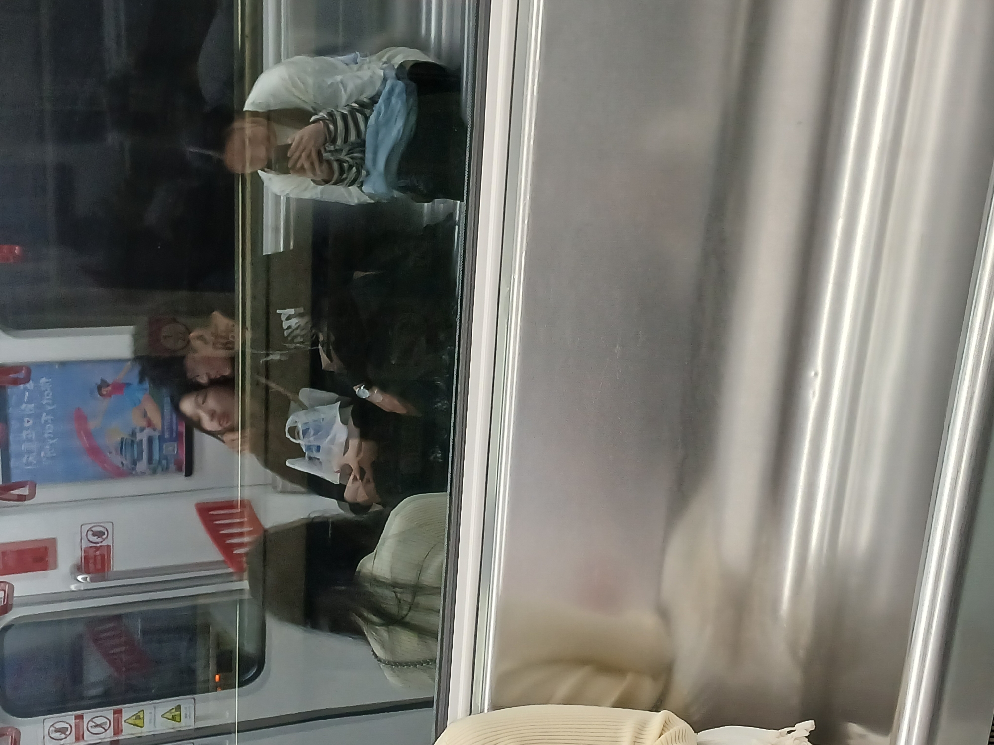The image depicts the interior of a train carriage, likely a subway or metro, in Chongqing High-tech Zone, China. The view is through a glass door, partially obscured by reflections. Several passengers are visible inside the carriage. The passengers appear to be resting or sitting quietly.
Noteworthy objects include the train's interior fixtures, posters, and signage. The general environment suggests an urban setting.