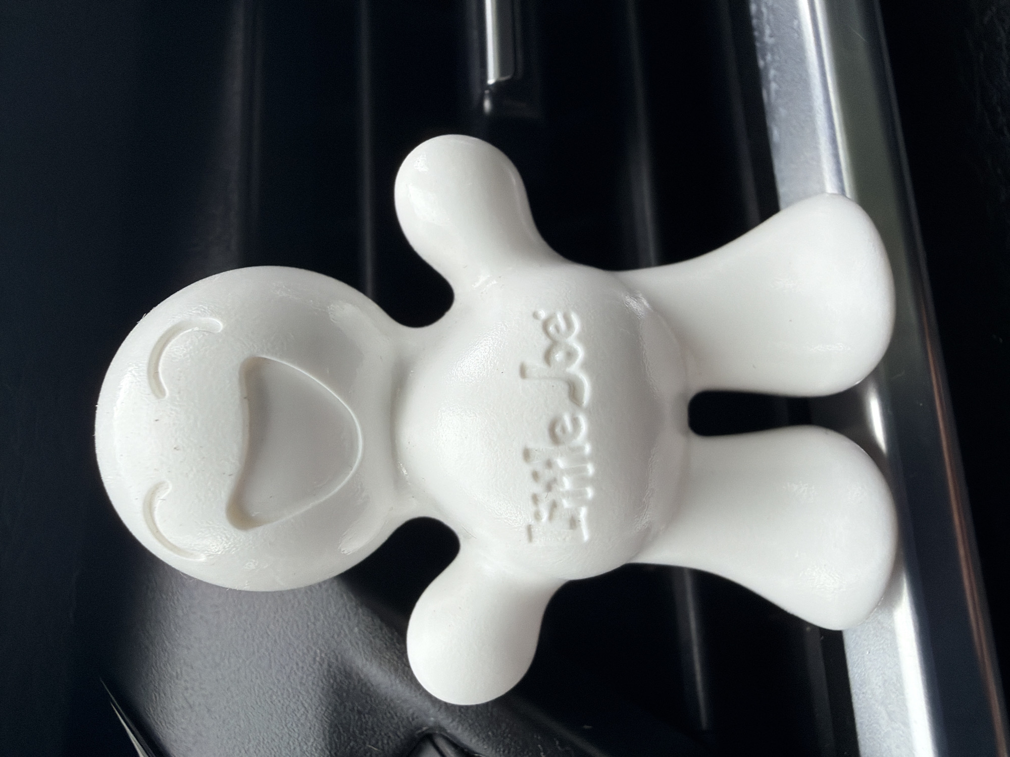 An air freshener shaped like a smiling cartoon character, branded "Little Joe," is affixed to a metallic car air vent. The character is white and roughly humanoid, with a large, open smile and curved eyes. The background is dark, suggesting the interior of a vehicle. The scene is illuminated by natural light, casting subtle shadows. The location is confirmed as Saurieši, Latvia.