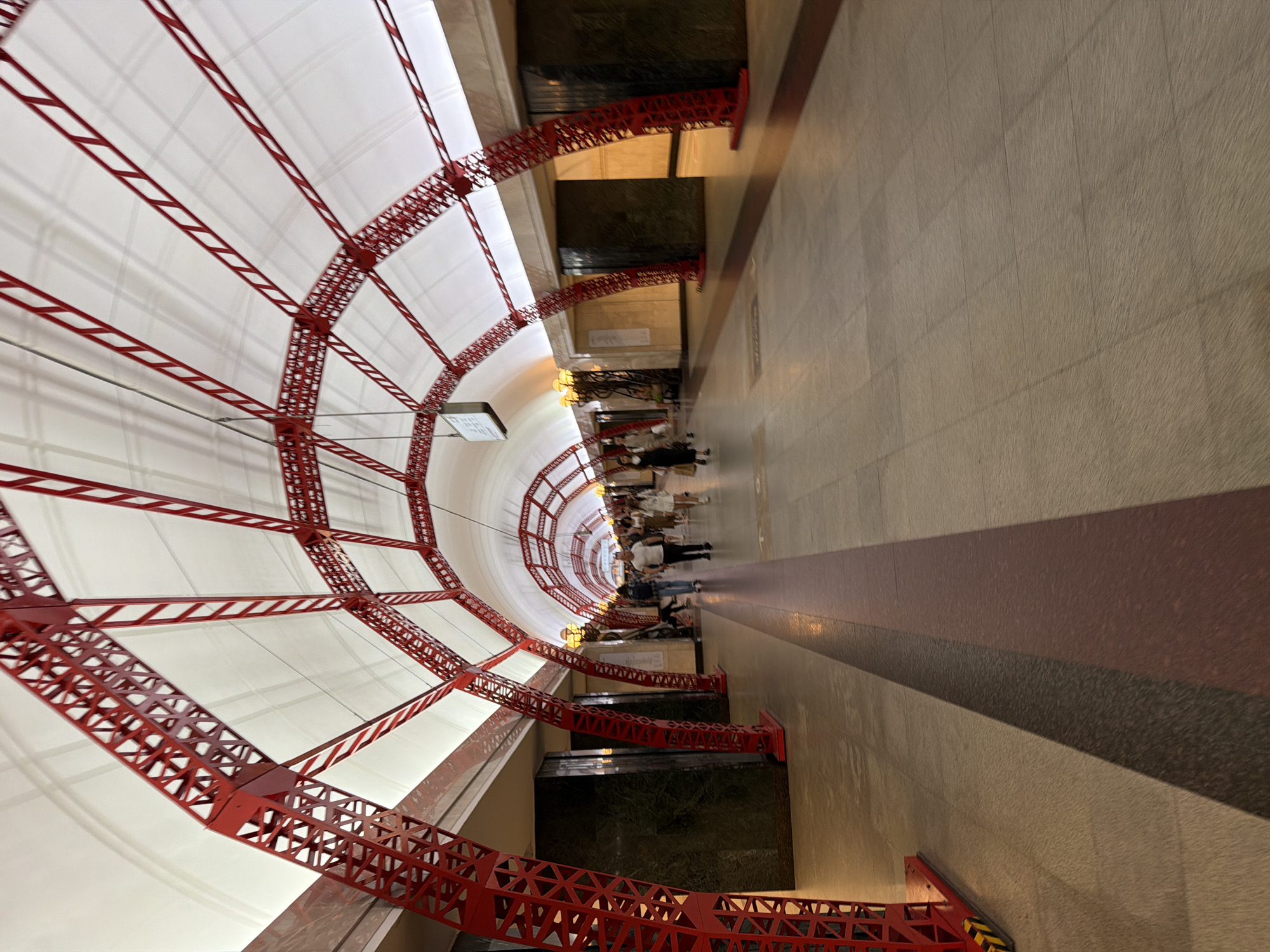 The image depicts a view looking down a long, arched subway station hallway. The ceiling is covered with a white, translucent material supported by a red, lattice-like metal structure that forms a series of arches. The floor is tiled in a neutral tone with darker, strip-like inlays. Several people are walking down the hallway in the distance, mostly appearing as silhouettes against the bright lighting further ahead. The walls of the station are lined with dark, polished stone and some lighter colored panels. The overall impression is one of an interior architectural space, likely a subway or metro station, with a distinct and decorative ceiling design.