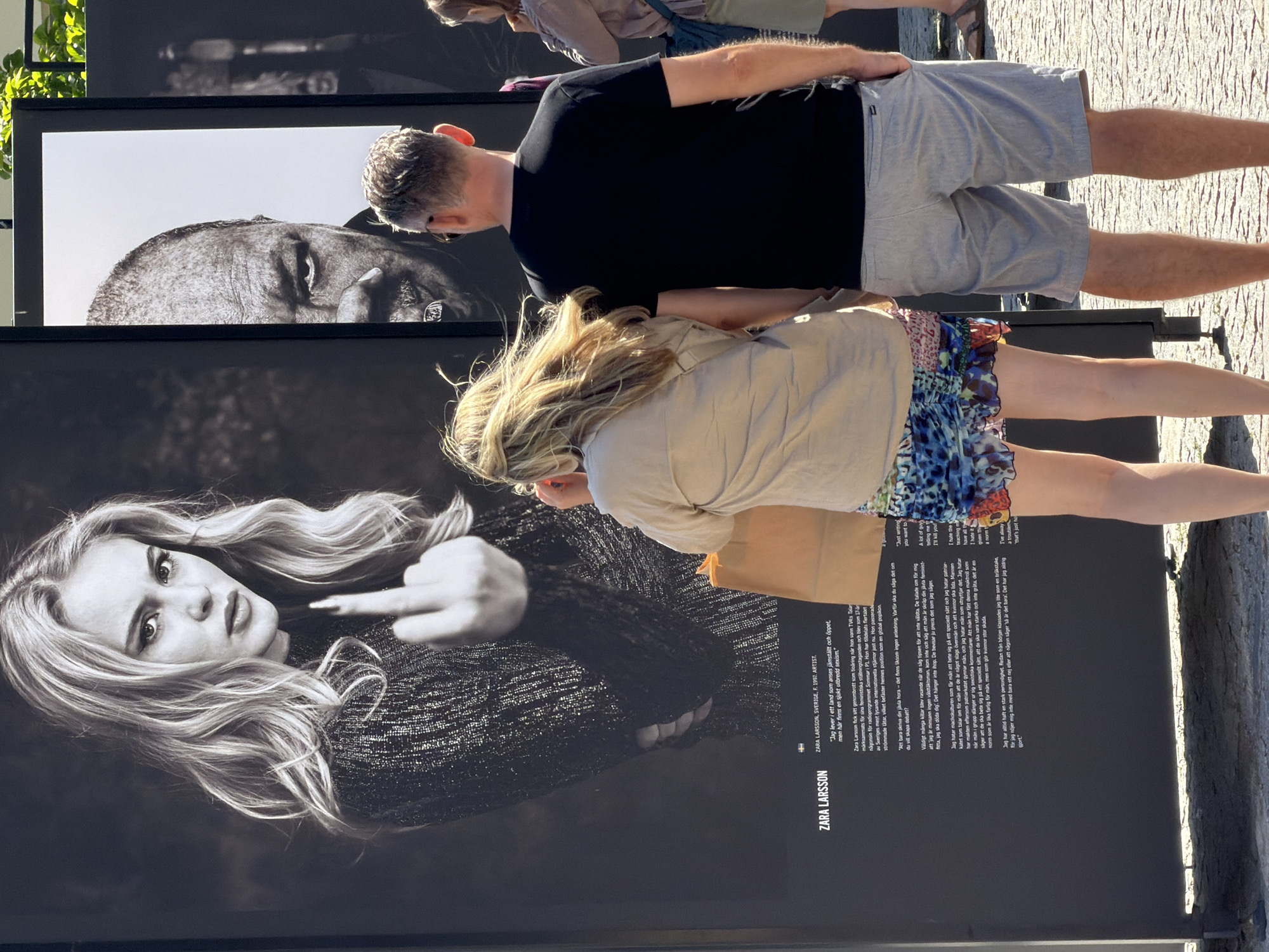 The image shows an outdoor exhibition in Lund, Sweden, with large black and white photographs displayed on panels. In the foreground, a woman wearing a beige shirt and a colorful patterned skirt stands with her back to the camera, holding a brown paper bag. Next to her, a man wearing a black t-shirt and light grey shorts also stands with his back to the camera, with his right hand in his pocket. Behind them, the photographs are of Zara Larsson, a Swedish singer and songwriter, and another individual, possibly a man, with his face partially visible. The Zara Larsson photograph shows her from the chest up, looking directly at the camera and holding up her middle finger. The text accompanying the Zara Larsson portrait provides biographical information about her, stating she is from Sweden, born in 1997, and is an artist. It also includes Swedish text which appears to be a quote from her. The exhibition appears to be set up on a paved surface, with hints of greenery and sunlight suggesting an outdoor, possibly urban, location. The overall impression is that of a public art display.