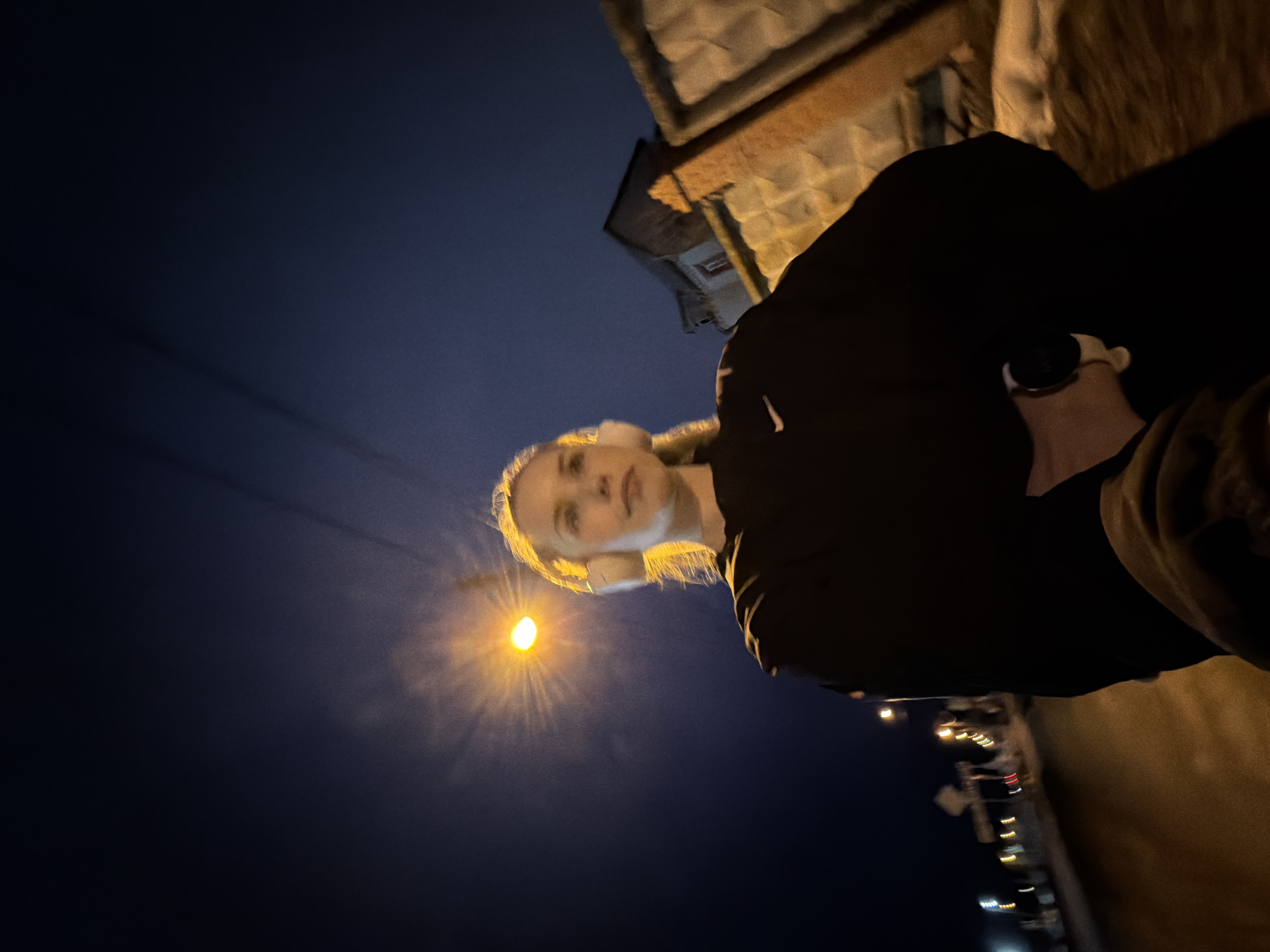 A low-angle, medium shot depicts a person standing on a street at night in Berezniki, Russia. The person has fair skin and blond hair and is wearing a black top. A streetlight shines brightly in the background, illuminating the person's face. In the background, a building and the street lights of a road can be seen.