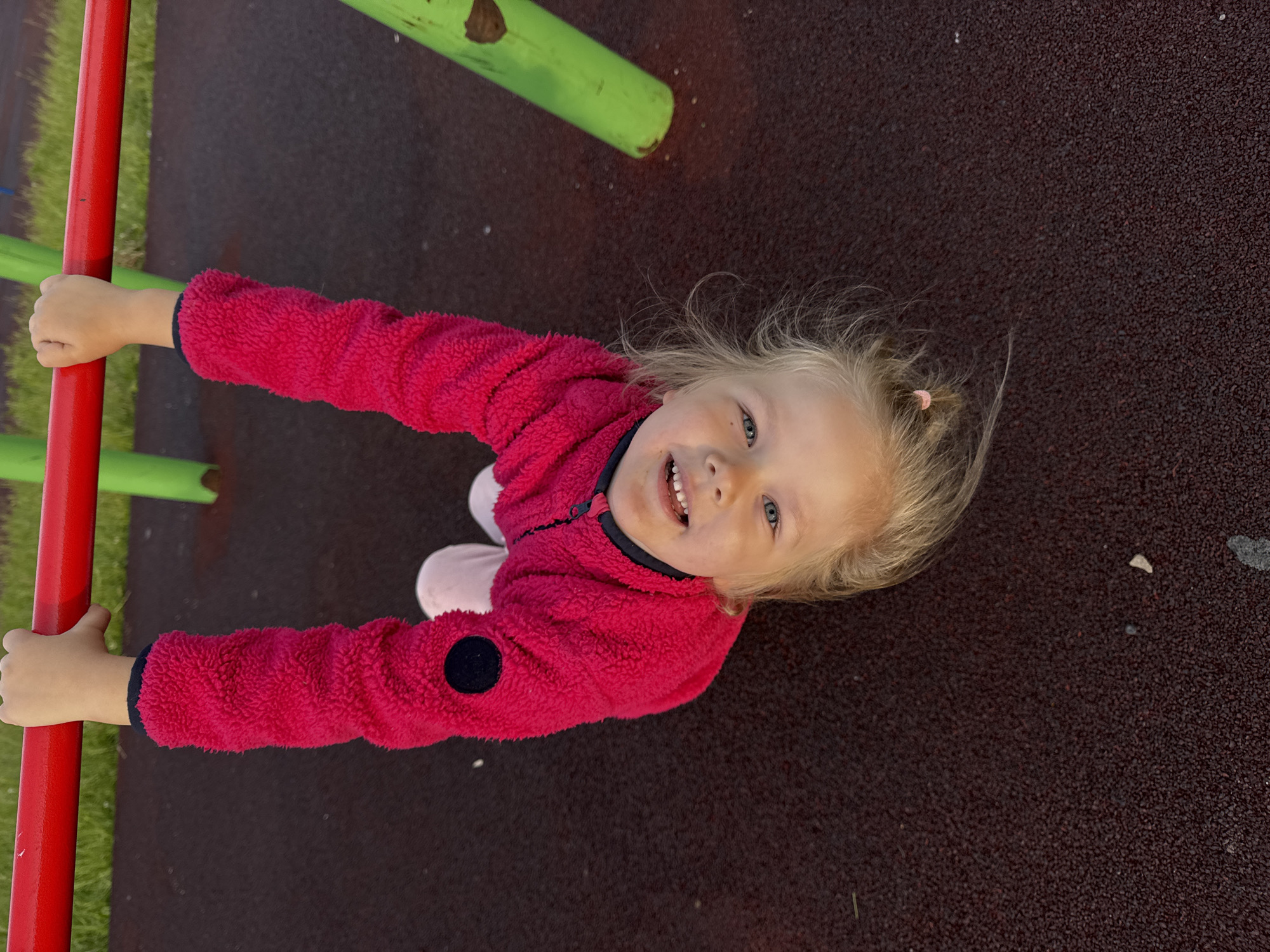 The image displays an outdoor playground scene in Горный, Russia, featuring a child approximately three to five years old hanging upside down from a red horizontal monkey bar. The child is wearing a fuchsia-pink textured fleece jacket with a visible black zipper pull and a black circular patch on the upper left sleeve. Light pink pants are also visible. Both hands are gripping the red bar above their head. The child has blonde hair and blue eyes, and is looking directly at the camera with an open-mouthed smile, revealing upper teeth. The ground beneath the child is a dark reddish-brown granular rubber safety surface. In the background, green metal support poles for playground equipment and areas of green grass are visible.