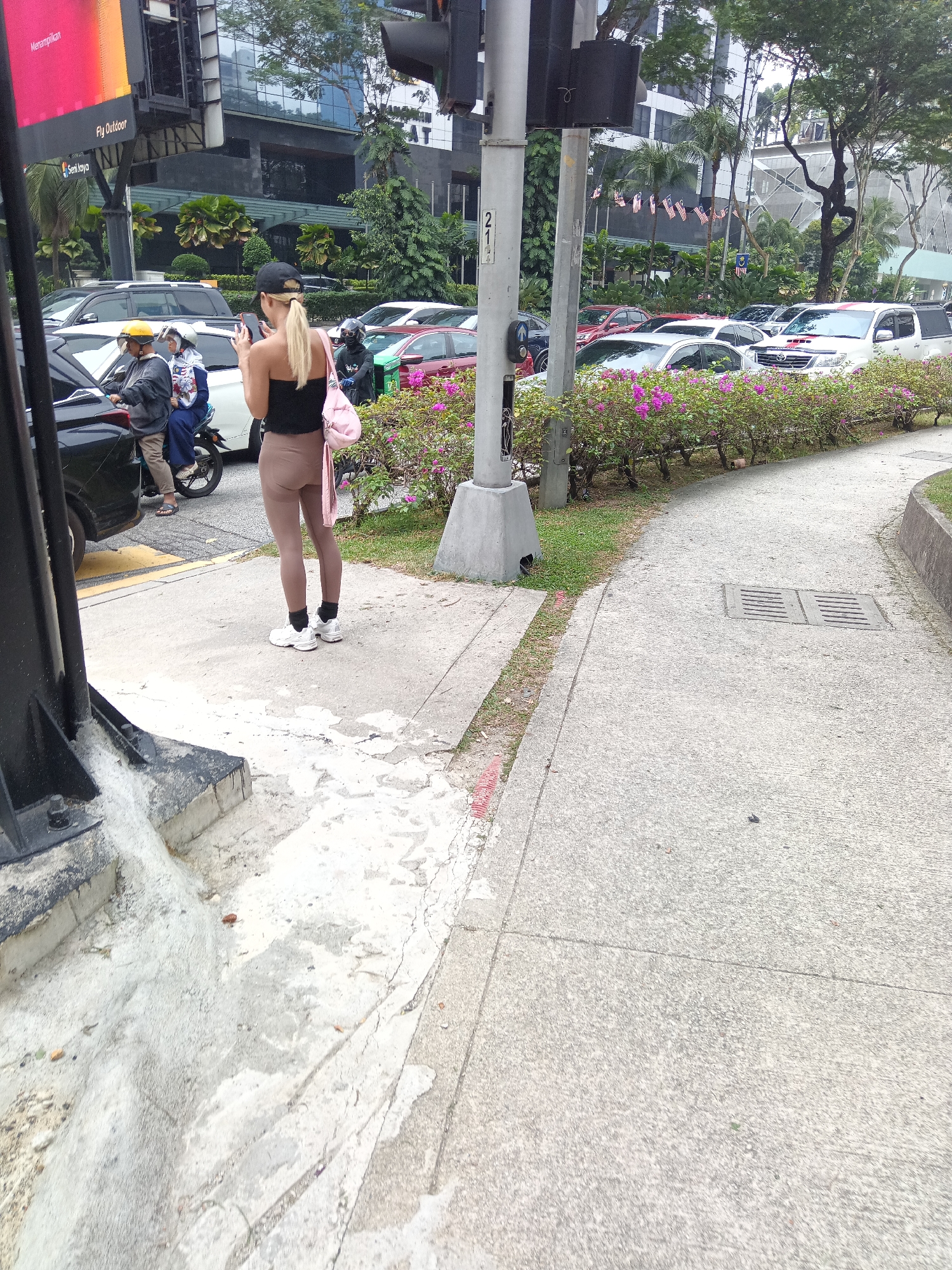 In Kuala Lumpur, Malaysia, on a cloudy day, a woman with blonde hair tied in a ponytail, wearing a black strapless top, tan leggings, and white athletic shoes, stands on a concrete sidewalk. She is wearing a black baseball cap and holding a pink purse over her left shoulder. She is looking down at her mobile phone in her hands. She is positioned near a traffic light and a street lamp. In the background, traffic is visible, including cars, motorcycles with riders wearing helmets, and a white SUV. Buildings and lush green trees line the street. The sidewalk is cracked and weathered in places, with some patches of grass and a drain grate visible. A curved pathway leads away from the foreground.