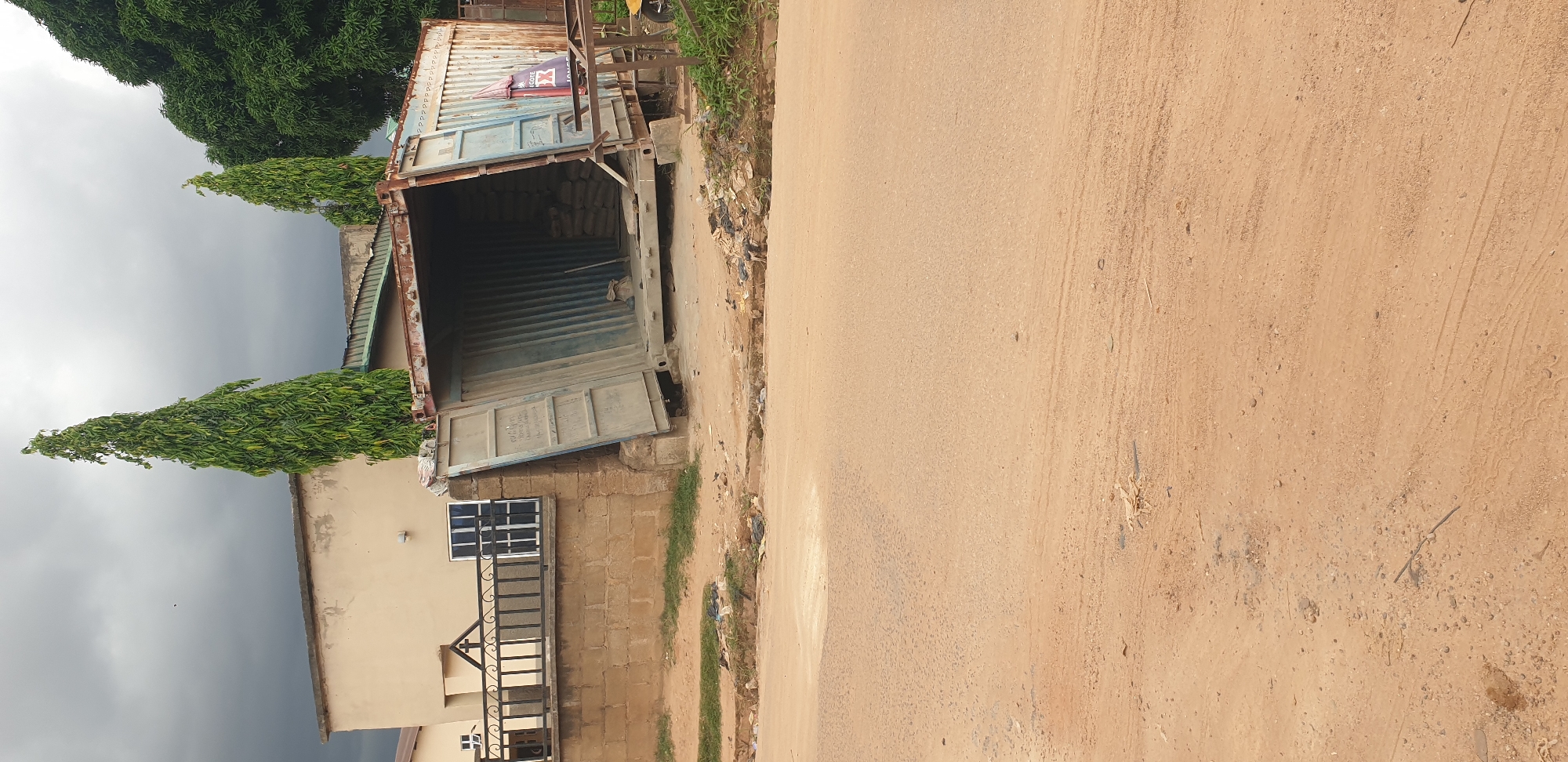 The image displays a roadside scene in Rido, Nigeria, under an overcast, grey sky. A large, heavily rusted blue shipping container with its double doors open stands prominently on the left side. Inside the container, stacks of bags, possibly containing cement or other bulk materials, are visible, suggesting it serves as a storage unit or a makeshift vendor stall.
To the left of the container is a light-colored building with a dark roof and a window with multiple dark panes. In front of the building, a low brick wall supports a black metal gate or fence. Several tall, slender green trees with dense foliage rise into the cloudy sky, alongside a larger, bushier tree in the background.
The right side of the image features a wide, unpaved, reddish-brown dirt road that appears dusty and shows faint tire tracks. Along the edge of the road, adjacent to the container, are patches of green grass and scattered refuse.
On the exterior of the rusted shipping container, a faded, torn banner or poster is attached, displaying remnants of red and white graphics. Partially visible text on the banner includes "IGODE" and a brand logo that resembles a stylized 'X'. No people are present in the scene, and no specific activity is taking place at the moment of the photograph.