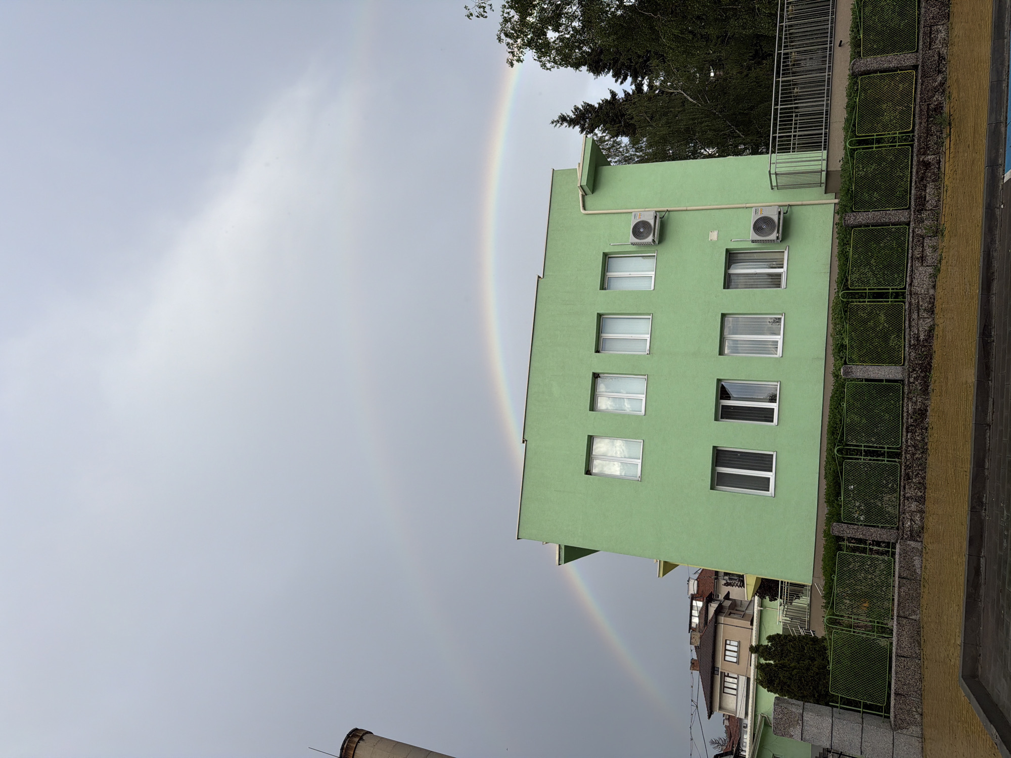 A rainbow arcs across the sky next to a green building with air conditioning units and white-framed windows. The building is located in Panagyurishte, Bulgaria. Part of the rainbow appears behind the building, touching the ground. The sky is overcast. Fencing and foliage are visible along the right edge. Other houses and trees are present. A smokestack is visible in the lower left.