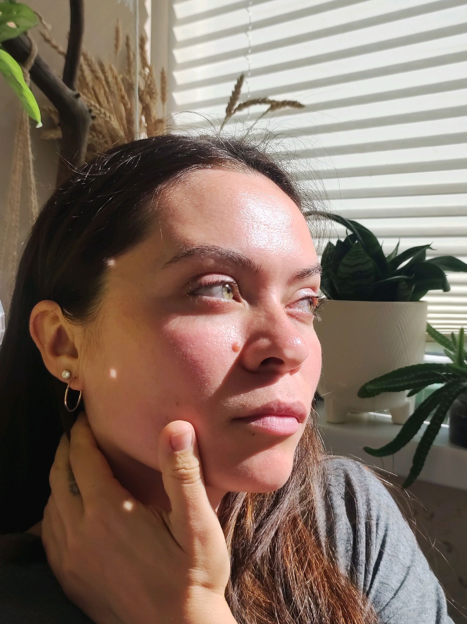 A woman, with her left hand touching her neck and chin, is photographed in profile against a background of white blinds and a window. Sunlight casts dappled shadows across her face, highlighting her skin and the texture of her hair. A potted snake plant and a smaller succulent are visible to her right, near the window. On her left ear, she wears a small stud earring and a thin hoop earring. Her dark brown hair falls loosely around her shoulders. The image appears to be an indoor portrait. There is no information provided to suggest the location is in Chaikovsky, Russia.