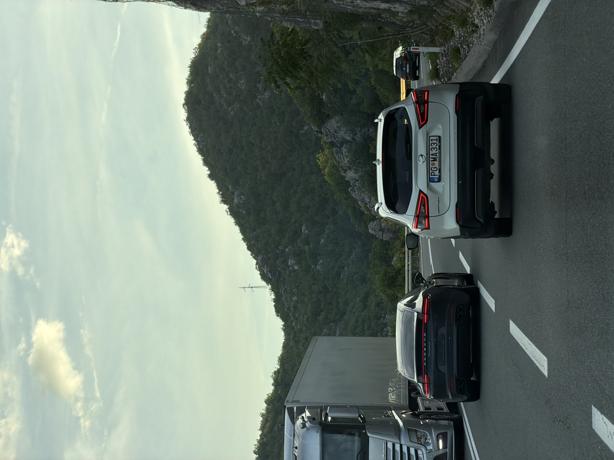 The image depicts a traffic scene on a road near Lipa Dobrska, Montenegro. A white Nissan Qashqai with license plate PG MA 331 is prominently visible, followed by a black Porsche Panamera and a truck. The surrounding environment features a mountainous landscape with trees and a partly cloudy sky. Overhead power lines are also noticeable.