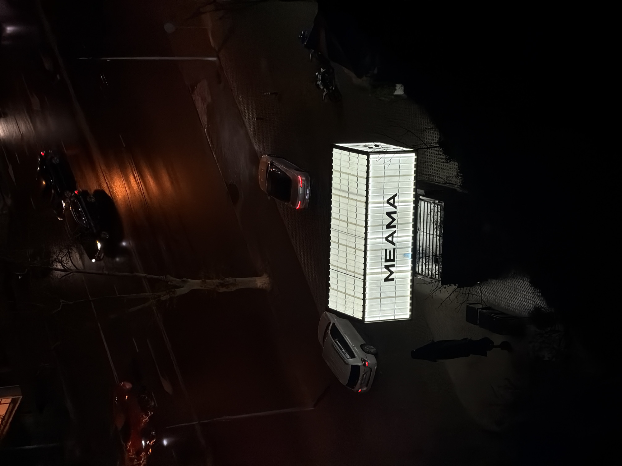 An aerial view at night shows a brightly illuminated building, identified by the large letters "MEAMA". The building is adjacent to a street where several vehicles are present, including parked cars. The surrounding environment is dark, with some light reflecting on the wet road surface. The location is Tbilisi, Georgia.