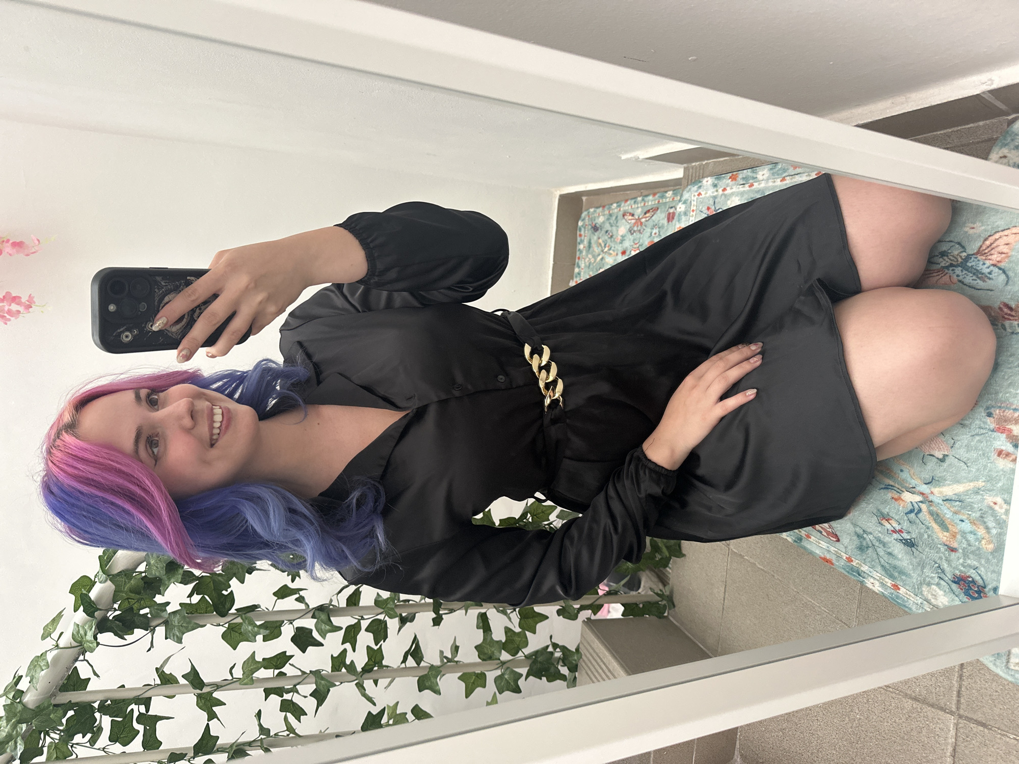 A young woman is captured in a mirror selfie, smiling brightly at the camera she holds in her right hand. Her hair is strikingly dyed in two distinct colors: vibrant pink on the left side of her face (viewer's left) and a deep blue or purple on the right. She has fair skin and her fingernails are adorned with glittery polish.
She is wearing a black, long-sleeved dress or tunic made of a satin-like material, featuring a collared neckline, buttons down the front, and puffy sleeves with elasticated cuffs. A prominent gold chain-link belt cinches her waist. The woman appears to be kneeling or squatting for the photo.
The setting is an indoor space, reflected in a full-length mirror. To the left of her reflection, a white decorative structure is visible, covered in green artificial ivy vines, and some delicate pink artificial flowers are also present. The floor visible in the reflection features light-colored tiles and a light blue patterned rug with motifs of butterflies and insects. The background walls are plain white. The lighting suggests it is daytime. No discernible text is visible. The scene is located in the City of Zagreb, Croatia.