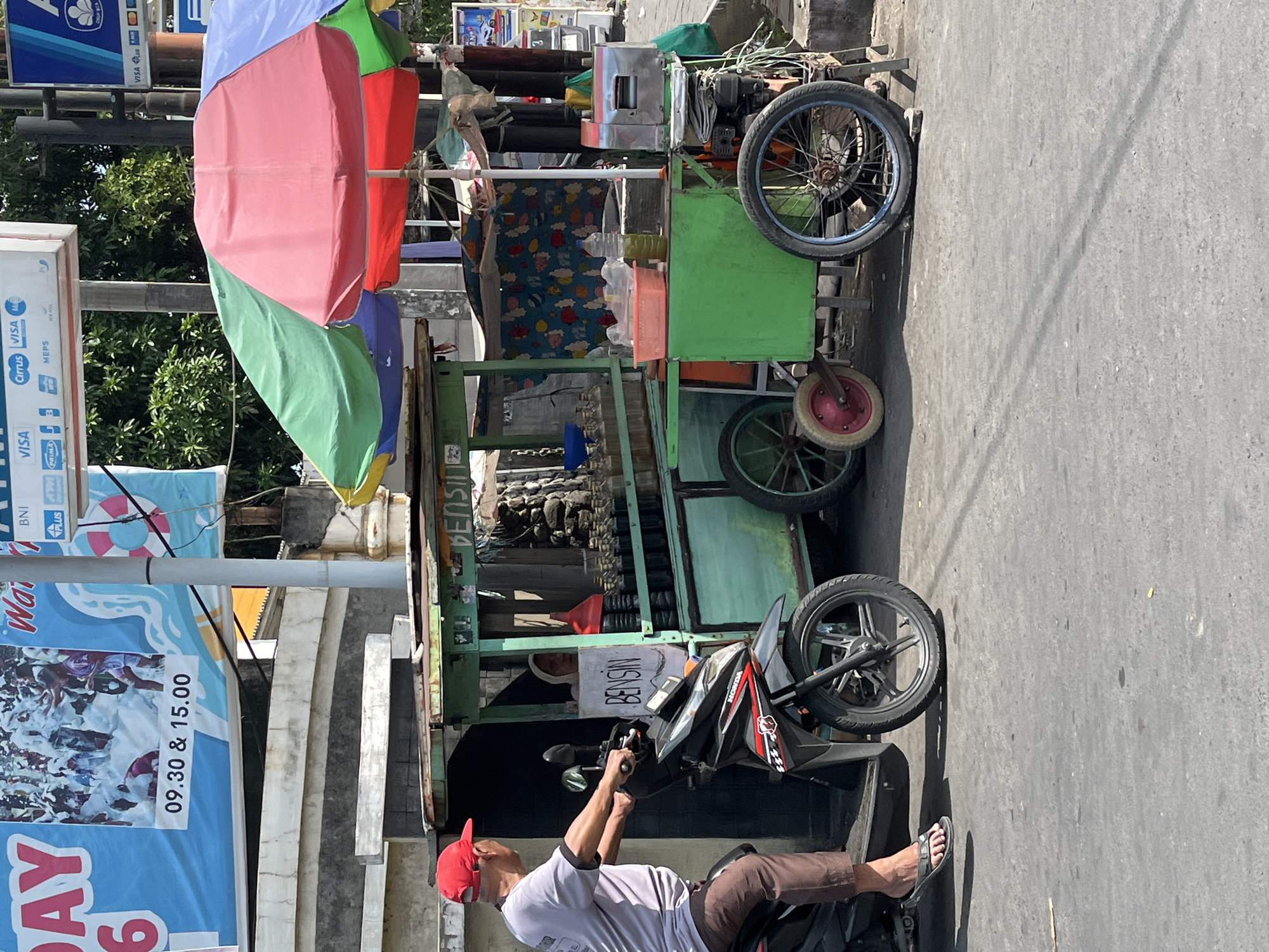 This is a medium shot, eye-level, full-color, outdoor photograph of a roadside scene in Boro, Indonesia. On the left, a man wearing a red baseball cap and brown pants rides a black and red Honda motorcycle. He is facing away from the camera. To the right of the motorcycle is a light green cart with multiple wheels, a green frame, and a red and green umbrella providing shade. The cart is selling what appears to be gasoline in bottles, with the word "BENSIN" written on a white sign in front of it. Behind the cart, a woman wearing a white hijab can be seen. A sugarcane juicer is also visible on the right side of the cart. In the background, there is a large blue banner with text and images, as well as trees and other structures. The ground is paved with asphalt.