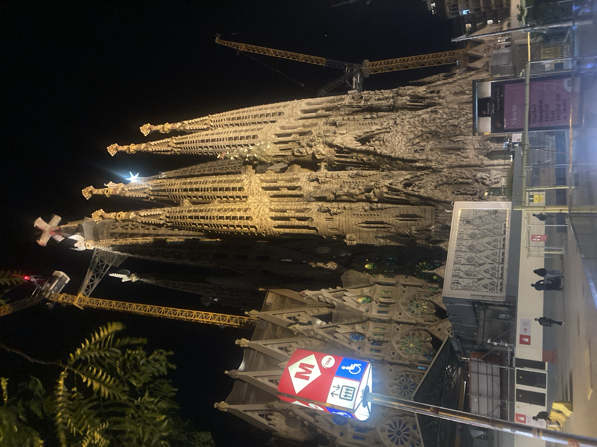 Here is a detailed description of the image:
The image depicts the Sagrada Familia cathedral in Barcelona, Spain, at night. The cathedral's ornate facade is illuminated and stands tall against the dark sky. Construction cranes are visible near the cathedral, indicating ongoing work on the building. A street sign with a large "M" is in the foreground, likely indicating a metro station. Pedestrians are walking on the sidewalk in front of the cathedral. A large banner is visible that advertises an art exhibit including digital art and the works of Warhol and Basquiat. Trees can be seen in the lower left corner. The overall impression is that of a bustling urban scene, with the Sagrada Familia as a prominent and iconic landmark.