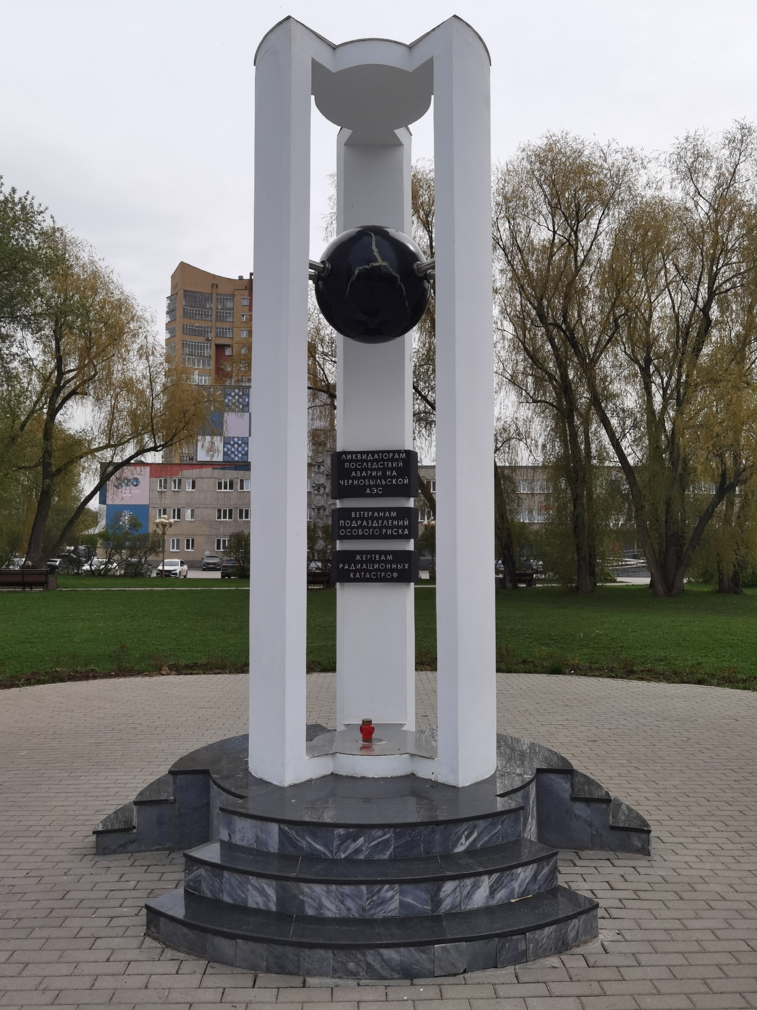 A medium shot captures a monument in Perm, Russia, set against a muted urban backdrop. The monument features a black sphere encircled by a white frame, standing on a tiered base of gray marble. A red votive candle rests on the monument's surface. Inscriptions in Cyrillic are visible on plaques affixed to the structure. The monument is situated in a park-like setting with paving stones underfoot and a grassy area surrounding the monument. Trees with sparse autumnal foliage flank the structure, and in the background stands an apartment building with a multi-colored facade. Cars are parked nearby, indicating an accessible urban environment. No people are visible in the immediate scene. The overcast sky suggests a cloudy day.