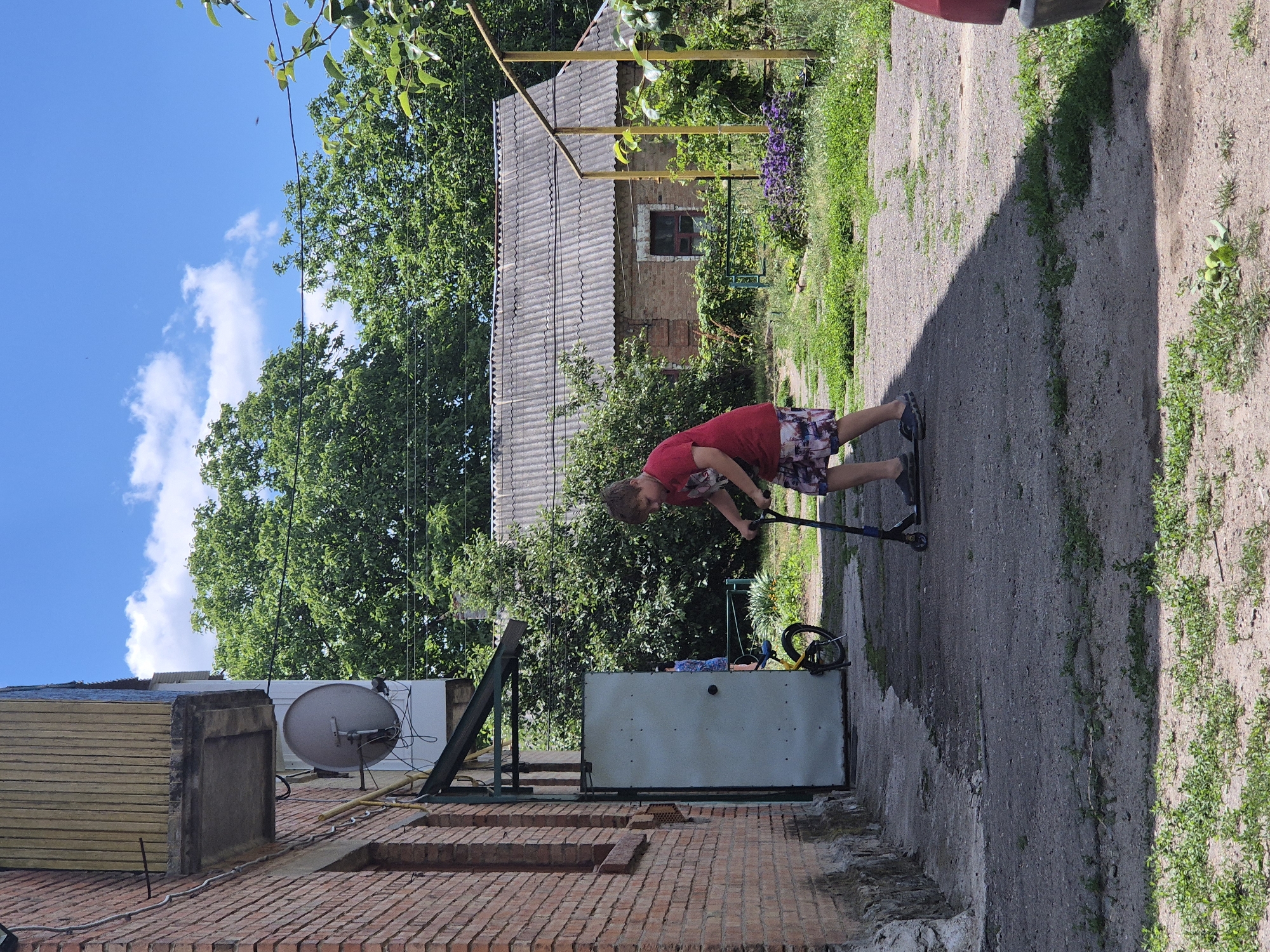 In Sozonivka, Ukraine, on a sunny day with a bright blue sky and scattered clouds, a young boy, approximately 10 years old, is riding a scooter on a gravel and concrete pathway next to a brick building. He is wearing a red t-shirt with a graphic print and multi-colored shorts. The building has a satellite dish mounted on its exterior and a metal awning over a large, light-colored door. To the right of the pathway is a yard with green grass, trees, and a low, gray-roofed building in the background. A yellow bicycle is leaning against the door. The scene suggests a residential area, possibly an apartment complex or private dwelling.
