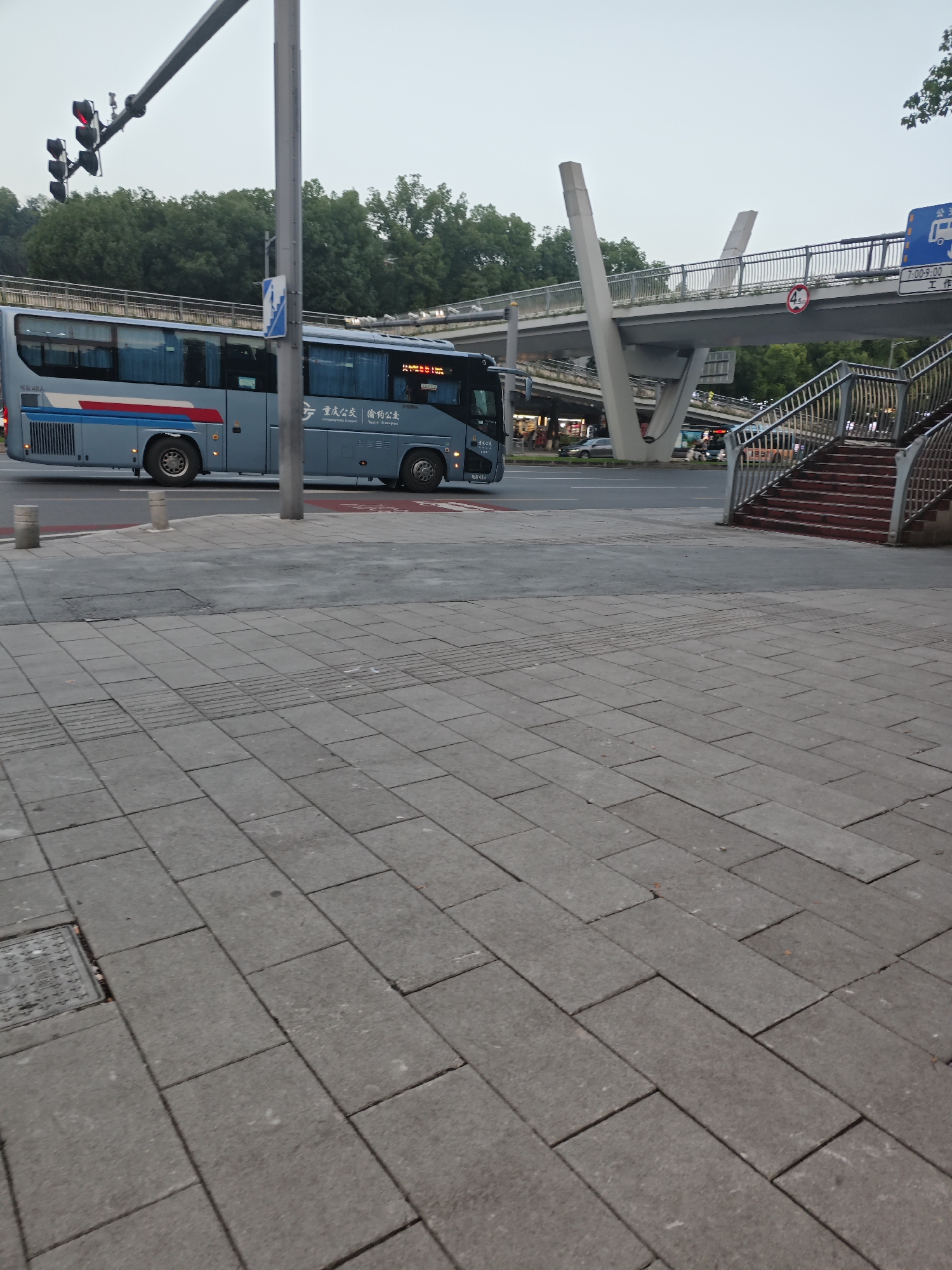 This image shows a street scene in the Chongqing High-tech Zone, China. The primary subject is a light blue bus, possibly a public transport bus, with red and blue stripes along its side. The bus is positioned on a road, suggesting it is in transit. In the background, there is an elevated pedestrian walkway or bridge supported by concrete pillars. The walkway has metal railings and stairs leading up to it. Traffic lights are visible on the left side of the image. The ground in the foreground is paved with grey rectangular tiles, characteristic of a sidewalk. The overall environment appears to be an urban setting with trees and other vegetation bordering the road and walkway. The sky is overcast, indicating potentially cloudy weather. There are no visible people in the immediate foreground, but some vehicles and possibly people can be seen on the road and on the pedestrian walkway in the background. The scene captures a moment of urban infrastructure and transportation.