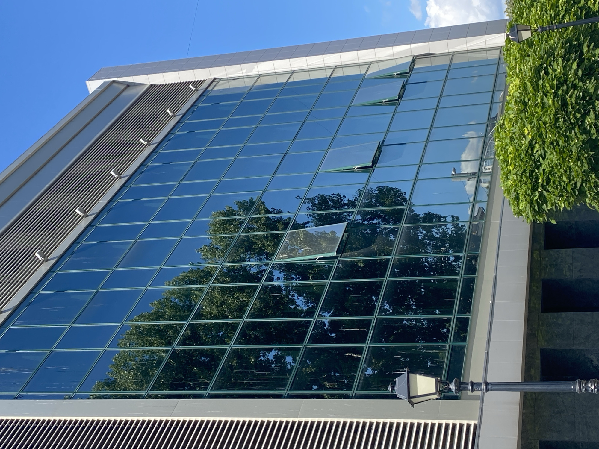 The image shows the facade of a modern building with large, reflective glass windows. The building has a light-colored exterior with horizontal louvers on the upper section. Several of the windows are open. A dark lamppost stands in front of the building on the left side, and lush green bushes are visible in the foreground. The sky is clear blue with some clouds. The reflection in the windows shows trees, the blue sky, and some buildings.
This specific building is located in Chisinau, Moldova, on Stefan cel Mare și Sfânt Boulevard, near the intersection with Tighina Street. This area is a central business district of the city.