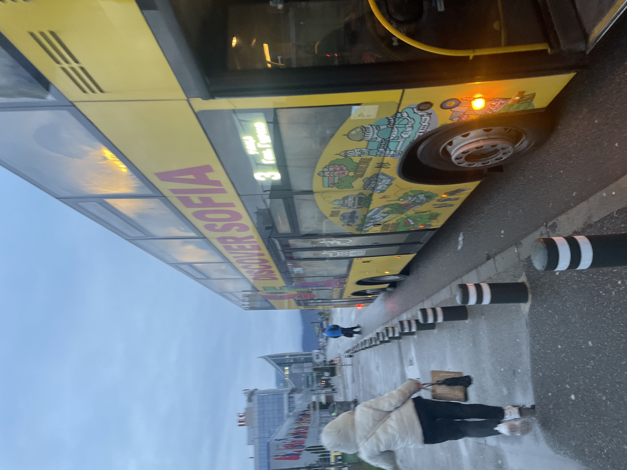 A slightly tilted medium shot depicts the side of a yellow "Discover Sofia" bus, parked along a sidewalk with black and white bollards. The bus features illustrations of Sofia landmarks and the number "3." A person wearing a white jacket and black pants walks on the sidewalk. In the background, buildings with flags are visible under a cloudy sky. The location is Sofia, Bulgaria.
