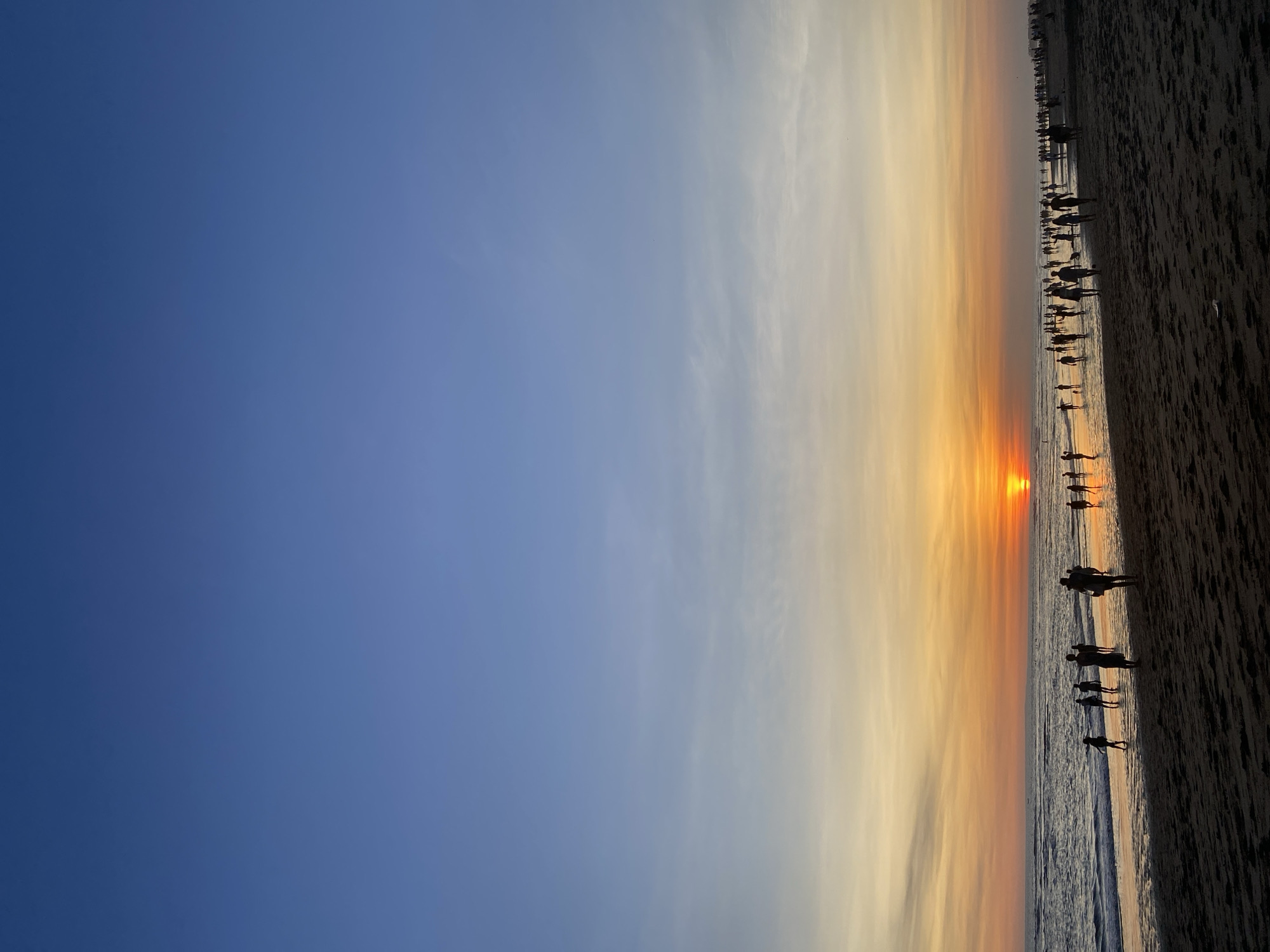 The image depicts a sunset scene on a beach. The sky transitions from deep blue to orange and yellow hues near the horizon. The sun is visible as a bright orange orb above the horizon line. A dark sandy beach occupies the bottom right portion of the image, with waves gently lapping the shore. Silhouettes of people are scattered along the beach and in the water, some standing and others walking. The scene is reminiscent of a beach in North Kuta, Indonesia during sunset.