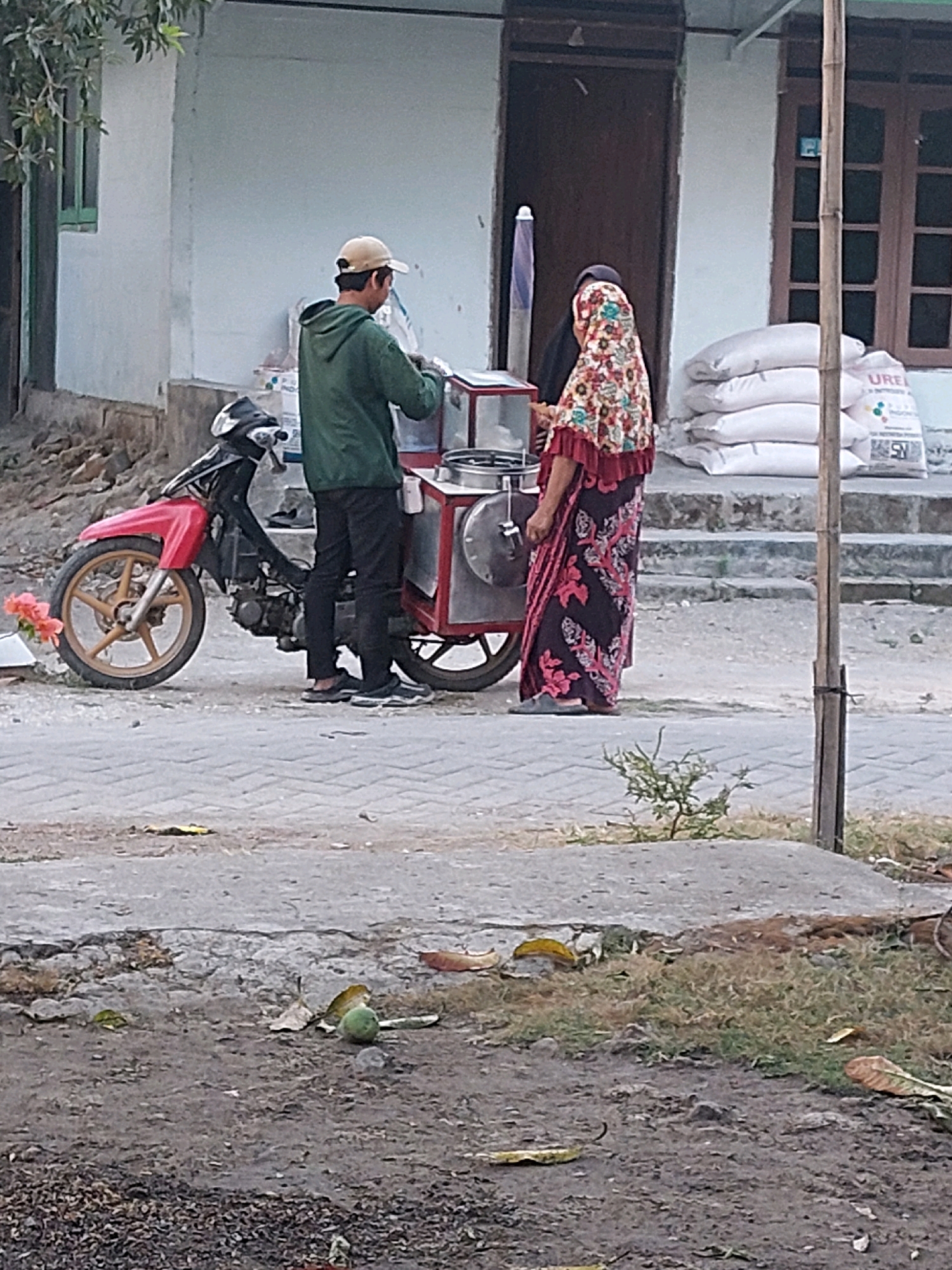 An outdoor scene in Sarangan, Indonesia features a street vendor with a mobile cart. A person wearing a green hoodie and a beige cap stands behind the cart, appearing to be serving a customer. To the right of the vendor, a woman wearing a colorful headscarf and a patterned dress is facing away from the camera, likely the customer. The cart is red and silver, with various containers and a metallic finish. Adjacent to the cart is a red and black motorcycle. In the background, a building with light blue walls and wooden-framed windows is visible. A stack of white sacks, some with visible labeling, are placed near the building's entrance. A bamboo pole stands vertically on the right side of the frame. The ground is paved with stone tiles in the foreground, with a dirt path and patches of dry grass further back. A single green fruit, possibly a mango, lies on the dirt. The overall setting suggests a commercial area or roadside stall.