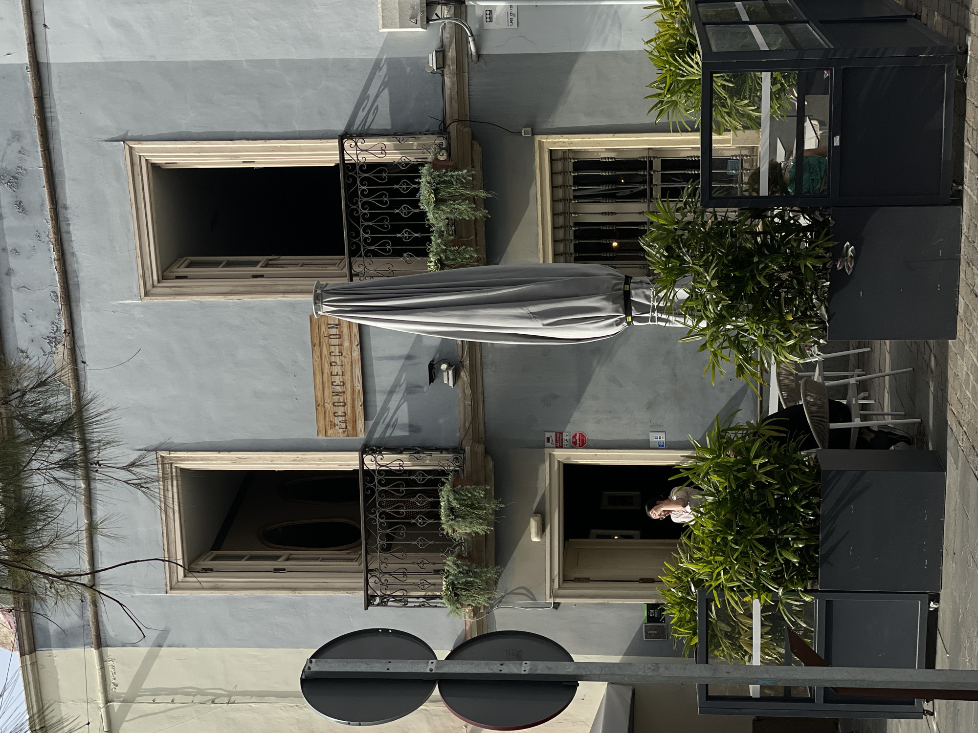 The image depicts the exterior of a building, possibly a restaurant, located in Santa Cruz de Tenerife, Spain. The building has a pale blue facade with two levels visible. The upper level features two windows, one with decorative wrought iron bars and flower boxes. A closed gray umbrella is mounted on the wall between the windows. A wooden sign with the words "LA CONCEPTION" is vertically displayed beside one of the windows. A person is visible inside the building's entrance on the lower level. The street is marked by street signs and large potted plants. Tables and chairs are set up outside the building.