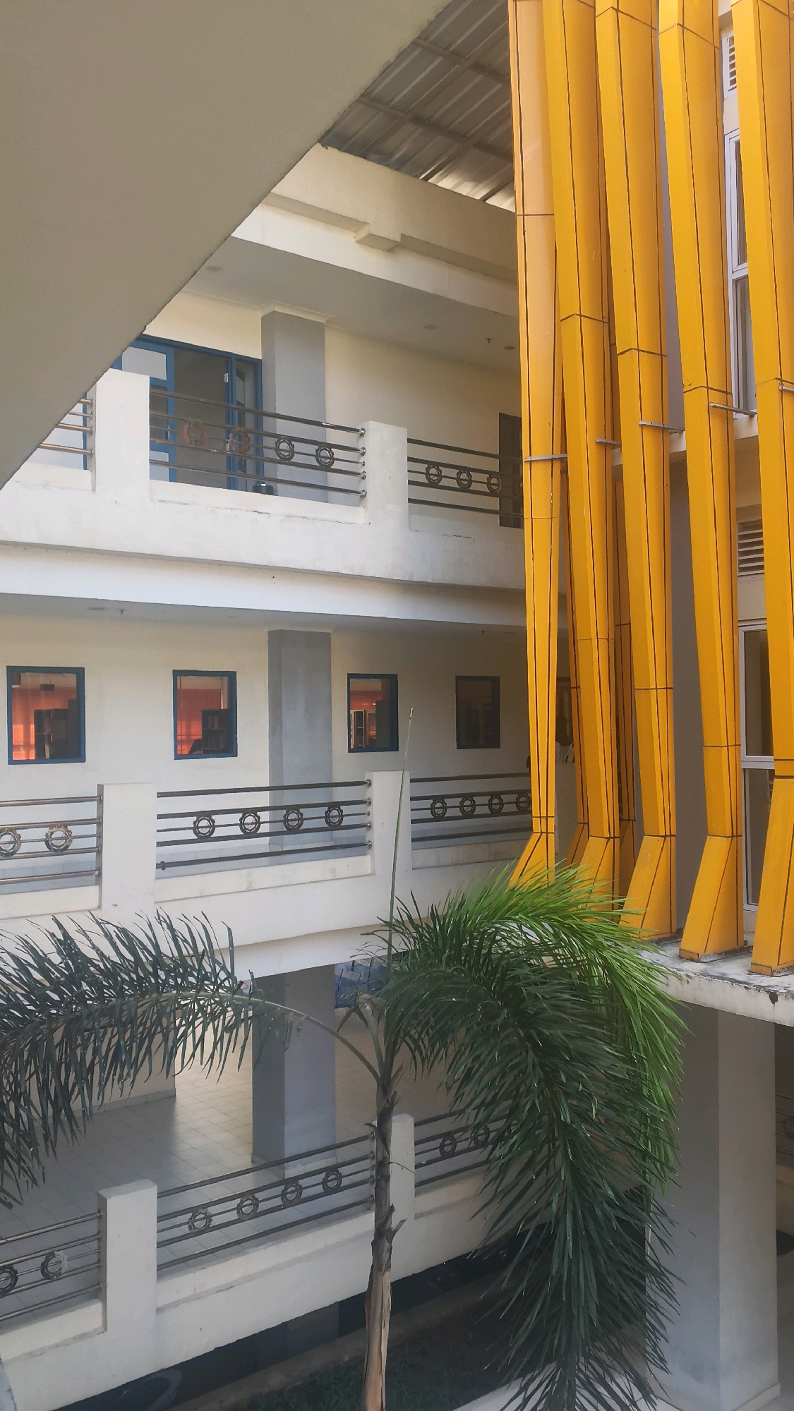 The image displays an outdoor courtyard of a modern building in Lohbener, Indonesia. The architecture features multiple levels with white balconies and railings, illuminated by bright sunlight. A prominent element is a row of tall, bright yellow vertical beams on the right side of the frame, possibly part of the building's facade or a decorative structure. Several windows are visible on the white walls, reflecting the warm orange glow of the interior. A palm tree with lush green fronds stands in the foreground, adding a tropical element to the scene. The courtyard appears to be an open space, with tiled flooring visible in the background. The overall impression is one of contemporary design within a bright, possibly warm climate.