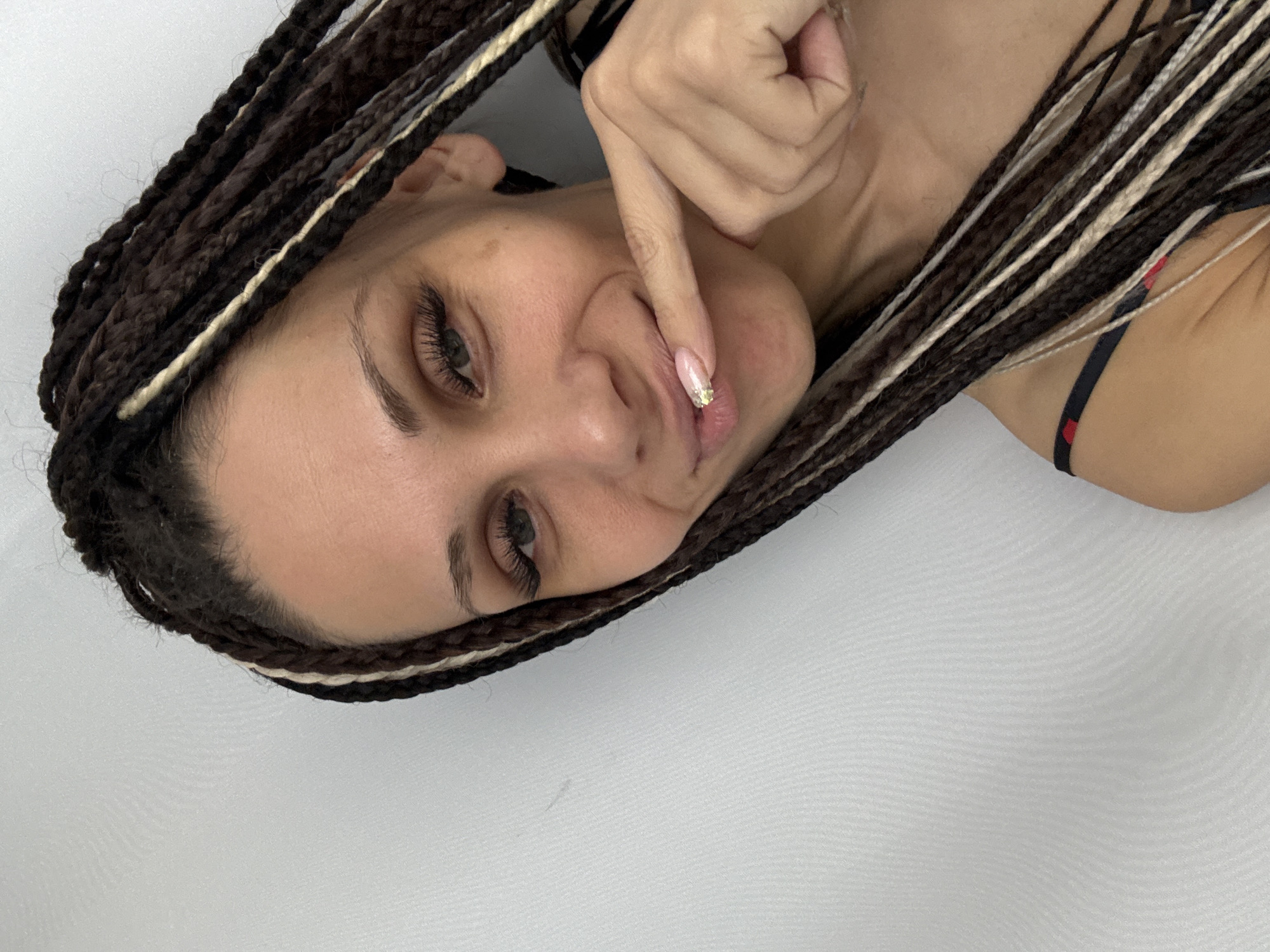 A young woman is lying on a white surface, possibly a bed or studio backdrop. Her head is surrounded by dark brown and white braids. She is touching her lip with her finger, which has a decorated nail. She has long eyelashes. The setting's location in Rusanovo, Russia is not discernible from the image itself.