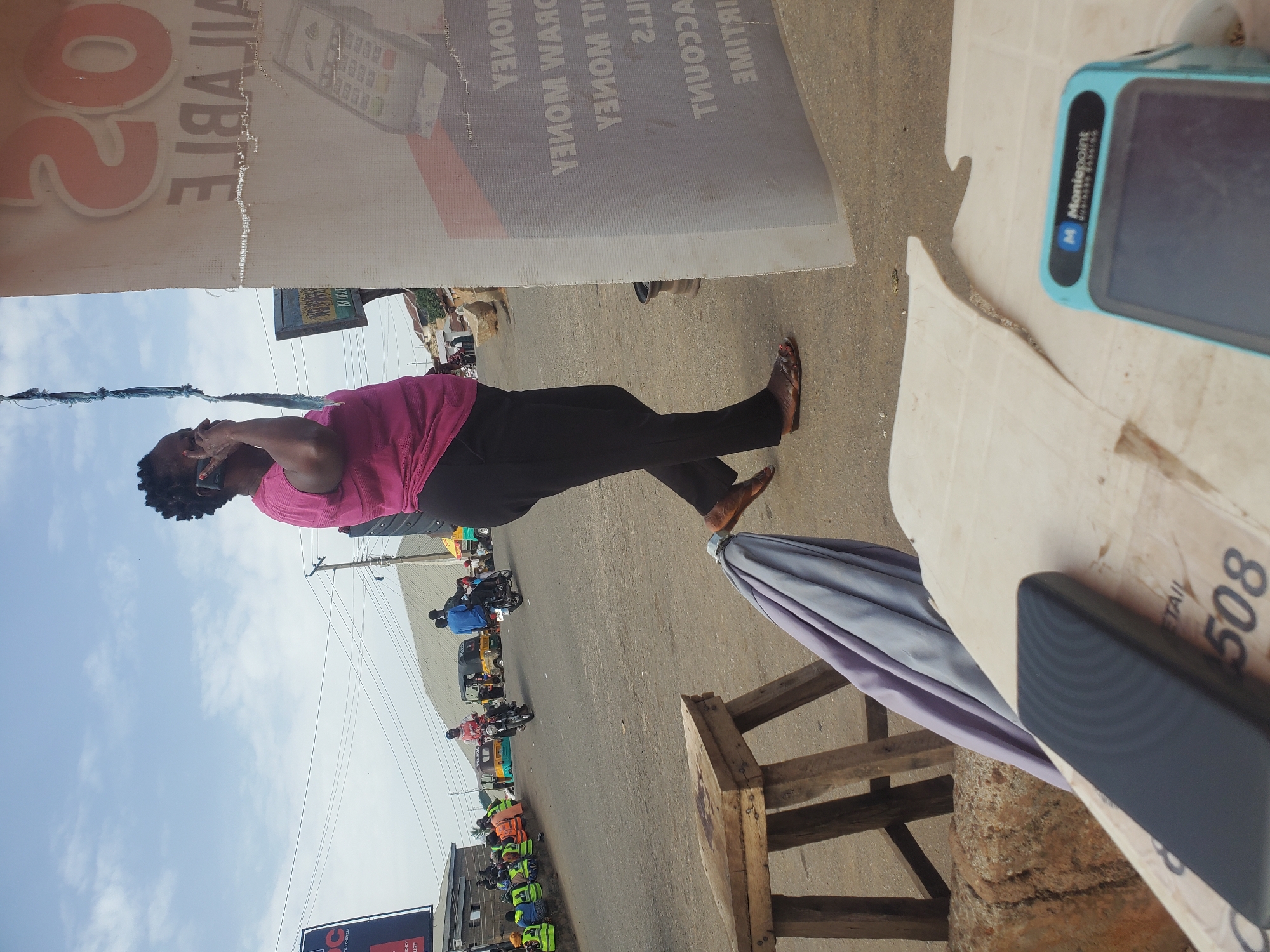A woman, with short, dark, braided hair, is standing on a dirt and asphalt road, talking on a cell phone. She is wearing a pink t-shirt and black pants. The woman is on the right side of the image, with the street and other people on the left. Behind her, a white banner with orange lettering is visible, advertising something with the word "AVAILABLE" clearly legible. The banner also depicts an image of a credit card machine. In the background, there are buildings, streetlights, and several motorbikes and a van on the road. A wooden stool is on the left side of the image, with a folded umbrella next to it. In the foreground, a light-colored surface, possibly a table, is covered with papers, one of which has the number "508" printed on it. A blue and black Moniepoint device is also visible in the foreground. The sky is overcast. The scene appears to be in a market or commercial area in Gwong, Nigeria.