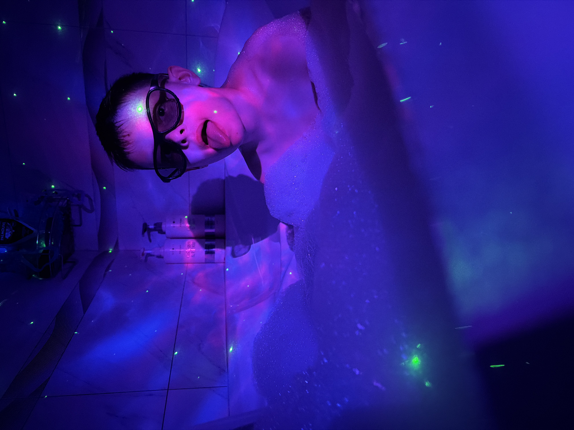 A person is submerged in a bubble bath in what appears to be a tiled bathroom, lit by a blue and purple light, possibly a blacklight. The person is wearing dark glasses and sticking their tongue out. Visible in the background are toiletry containers, and a bottle holder. The setting is a bathroom somewhere. There's no evidence in the photograph to place the location in Votkinsk, Russia.