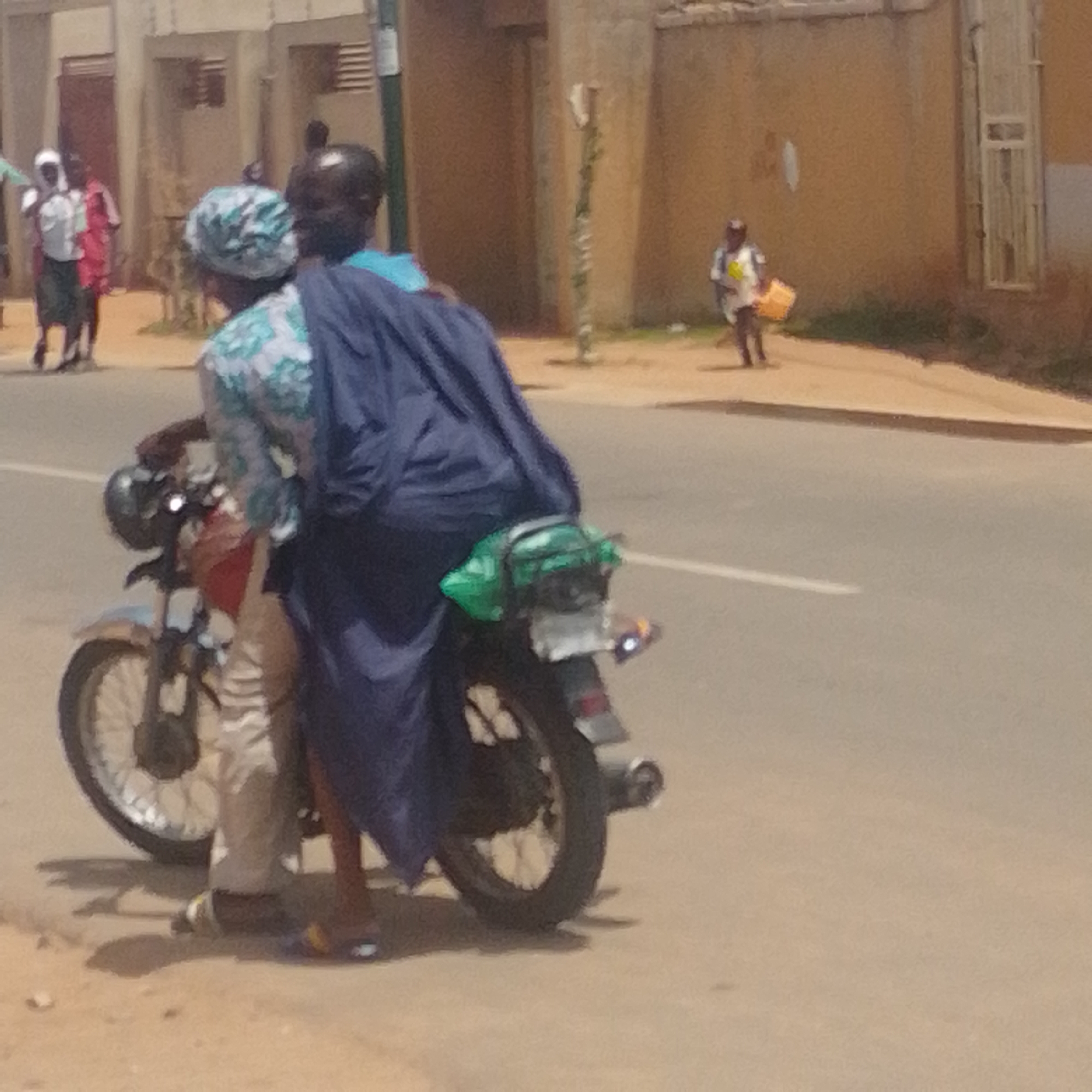 Two individuals are riding on a motorcycle in Gombe, Nigeria. The person in the front appears to be female, wearing a headscarf and a patterned top. The person in the back is wearing a dark blue garment that covers their lower body and the rear of the motorcycle. The motorcycle has a green bag strapped to the back. In the background, other people are visible walking along a street lined with buildings. One person in the distance is carrying a yellow container. The environment suggests an outdoor urban setting with a paved road and sidewalks.