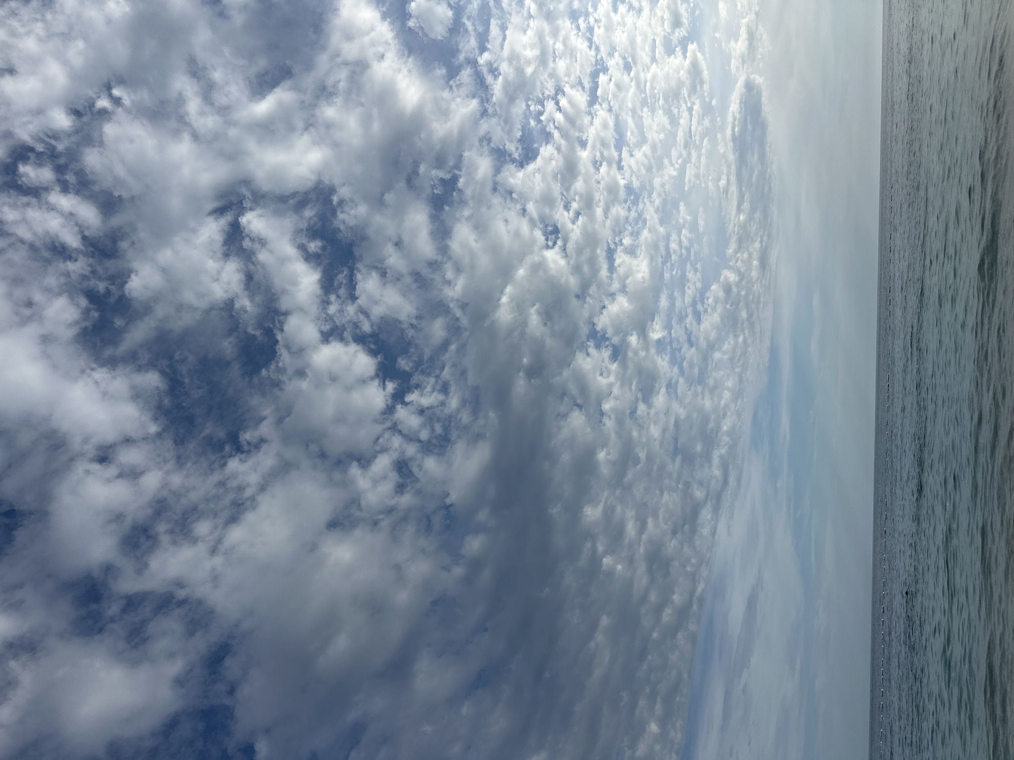 The image depicts a coastal scene in Mirište, Montenegro. The sky, occupying the upper two-thirds of the frame, is largely covered by a mixture of stratocumulus and altocumulus clouds; these clouds vary from dense, dark grey formations in the upper left to more scattered, whiter clouds against a lighter blue background in the lower right. A clear, horizontal horizon line separates the sky from the sea. The water is a calm, blue-green hue with subtle ripples on its surface. A line of white buoys extends horizontally across the mid-distance of the ocean. In the lower-mid left section of the water, a dark, partially submerged human-like figure is visible. The immediate foreground shows shallow, clear water with hints of the seabed.