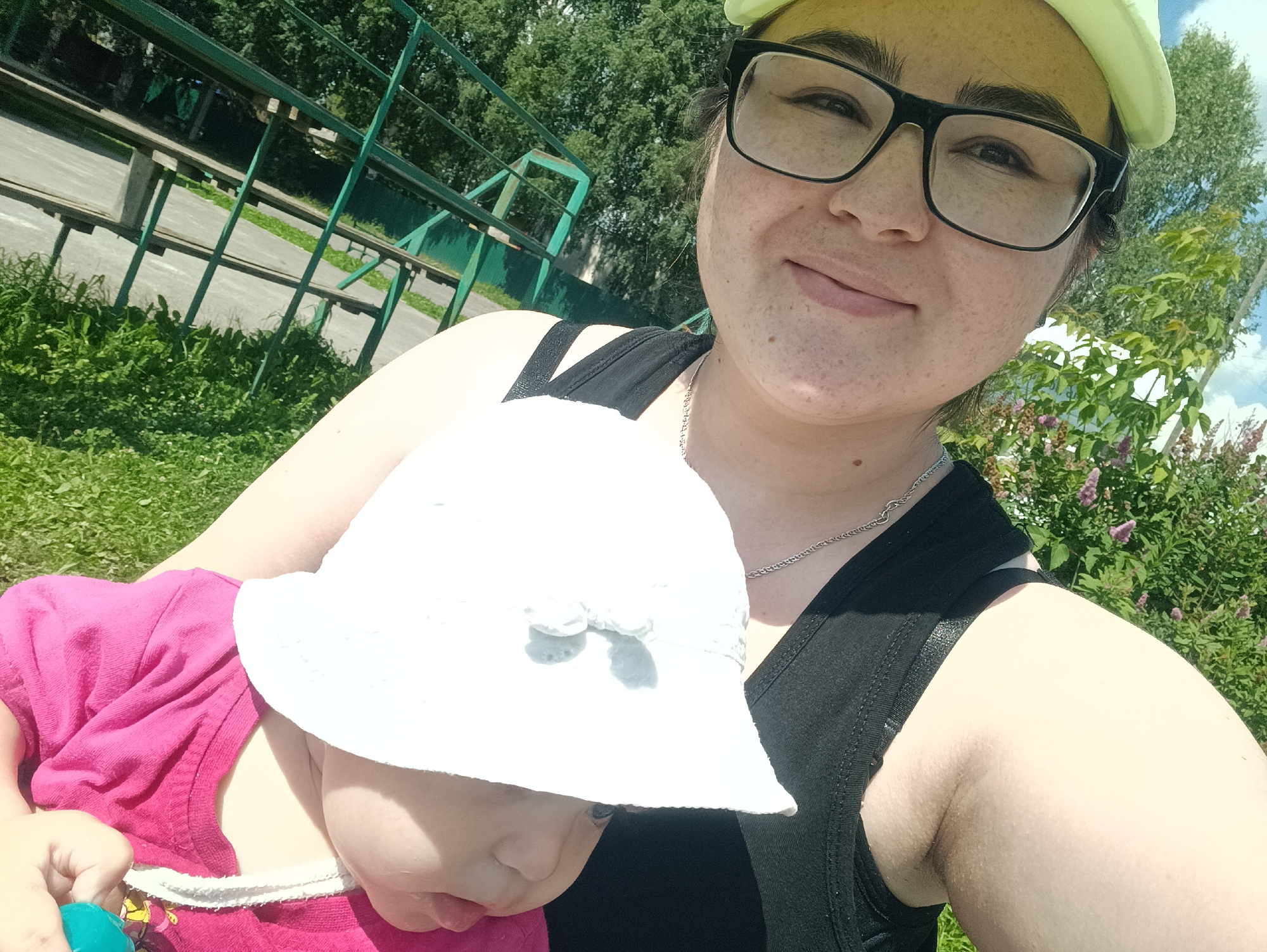 An outdoor, medium shot selfie shows a woman in a light yellow baseball cap and black tank top holding a baby wearing a pink shirt and a white bucket hat. The woman wears black-rimmed glasses and has light skin with freckles. She is smiling and looking at the camera. The baby is mostly obscured by the woman's arm and the white hat, but a portion of its face and torso are visible. The background features green grass, trees, and a portion of a green metal bleacher structure. The lighting suggests it is daytime and sunny.