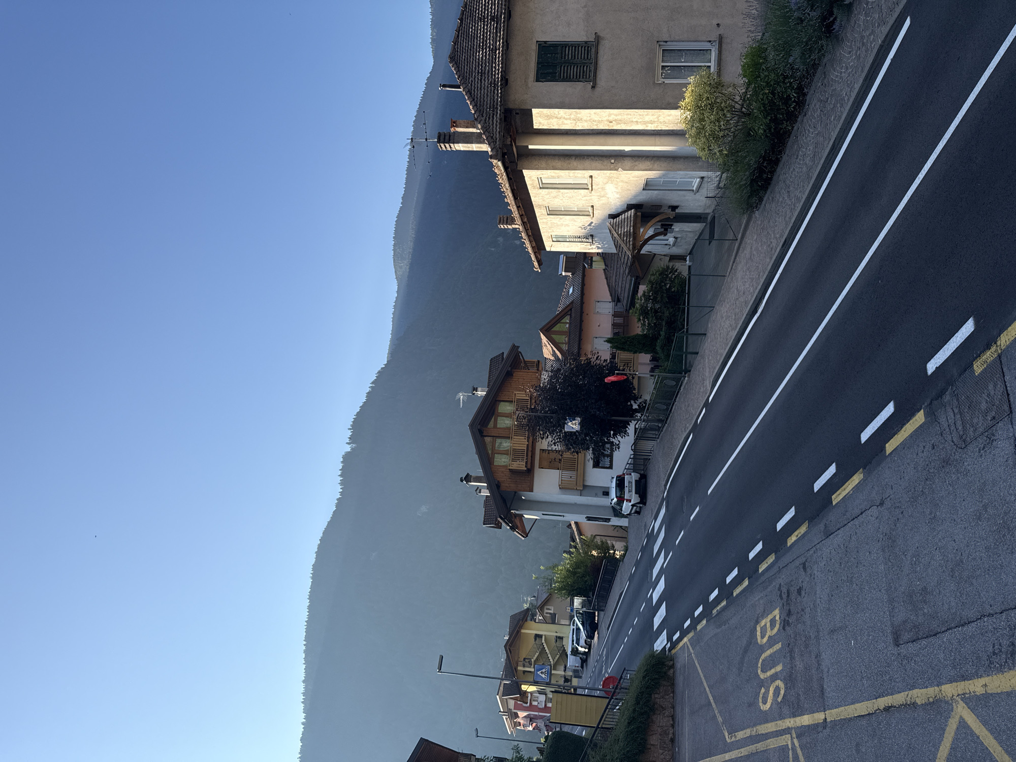 The image depicts a street scene in Molveno, Italy, under a clear blue sky. A sloping asphalt road with white lane markings dominates the foreground. To the left, a bus stop is marked with yellow lines and the word "BUS" painted on the ground. A crosswalk with white stripes is visible across the road. Several cars are parked along the street.
On the right side of the road, a light-colored building with a tiled roof and green shutters stands out. Other buildings, characteristic of Italian architecture with tiled roofs and balconies, are visible further up the street. A dense, green forested mountain range rises in the background, partially shrouded in mist. The scene appears to be captured in the morning or late afternoon, given the angle of the sunlight casting shadows. No people are actively engaged in any actions within the frame. The environment suggests a quiet, residential area with a backdrop of natural beauty.