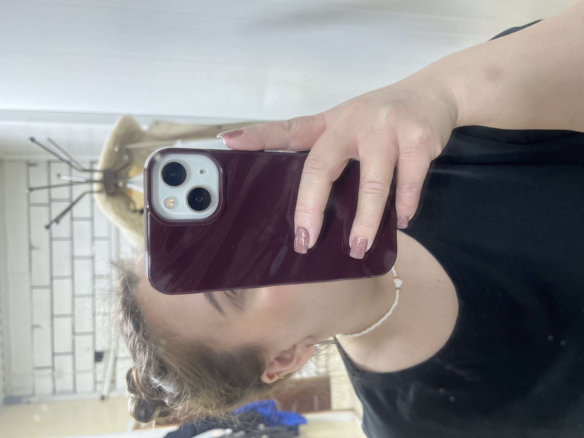 A woman is taking a selfie in a mirror, holding a phone with a dark red case. She is wearing a black shirt and a white beaded necklace. Her nails are painted with a glittery, light pink polish. The reflection in the mirror shows a chair, a coat rack, and a white wall. There is no definitive evidence within the image to suggest the location is Novopokrovskaya, Russia.