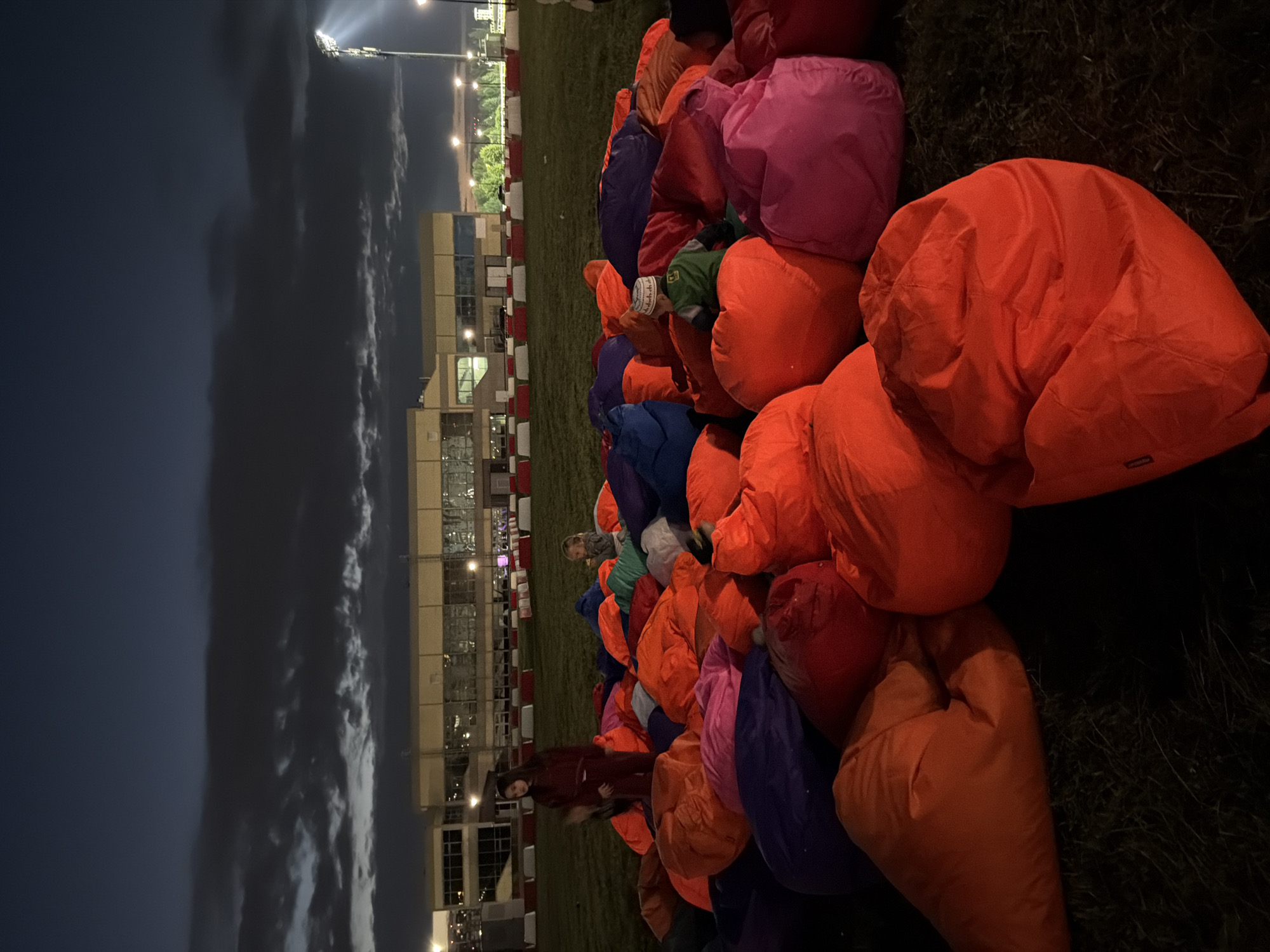 In the city of Unknown, Unknown, at night, a group of people are gathered on a grassy field, seated on a large pile of brightly colored beanbag chairs. The beanbag chairs are predominantly orange, with other colors like pink, purple, and blue visible. In the background, a modern building with large windows is illuminated, and beyond that, stadium lights are visible. The sky is dark with some clouds, and there are trees and fencing in the mid-ground. A young girl in a maroon tracksuit stands among the beanbag chairs, while other children are seated or playing. A young boy wearing a green jacket and a white and patterned cap is also visible. The overall scene suggests an outdoor event or gathering, possibly a movie screening or concert, set against an urban backdrop.