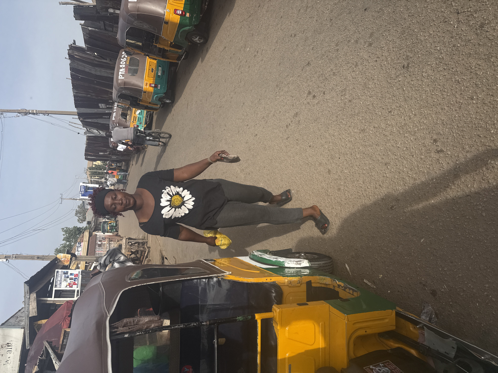 The image shows a woman standing in the street in Jos, Nigeria, next to a yellow and green tricycle taxi. She is wearing a black t-shirt with a white flower, gray pants, and sandals. She is holding a yellow rag and a small black object. Behind her are several more tricycle taxis parked along the road. In the background are shops and utility poles. A man on a bicycle is visible further down the street.