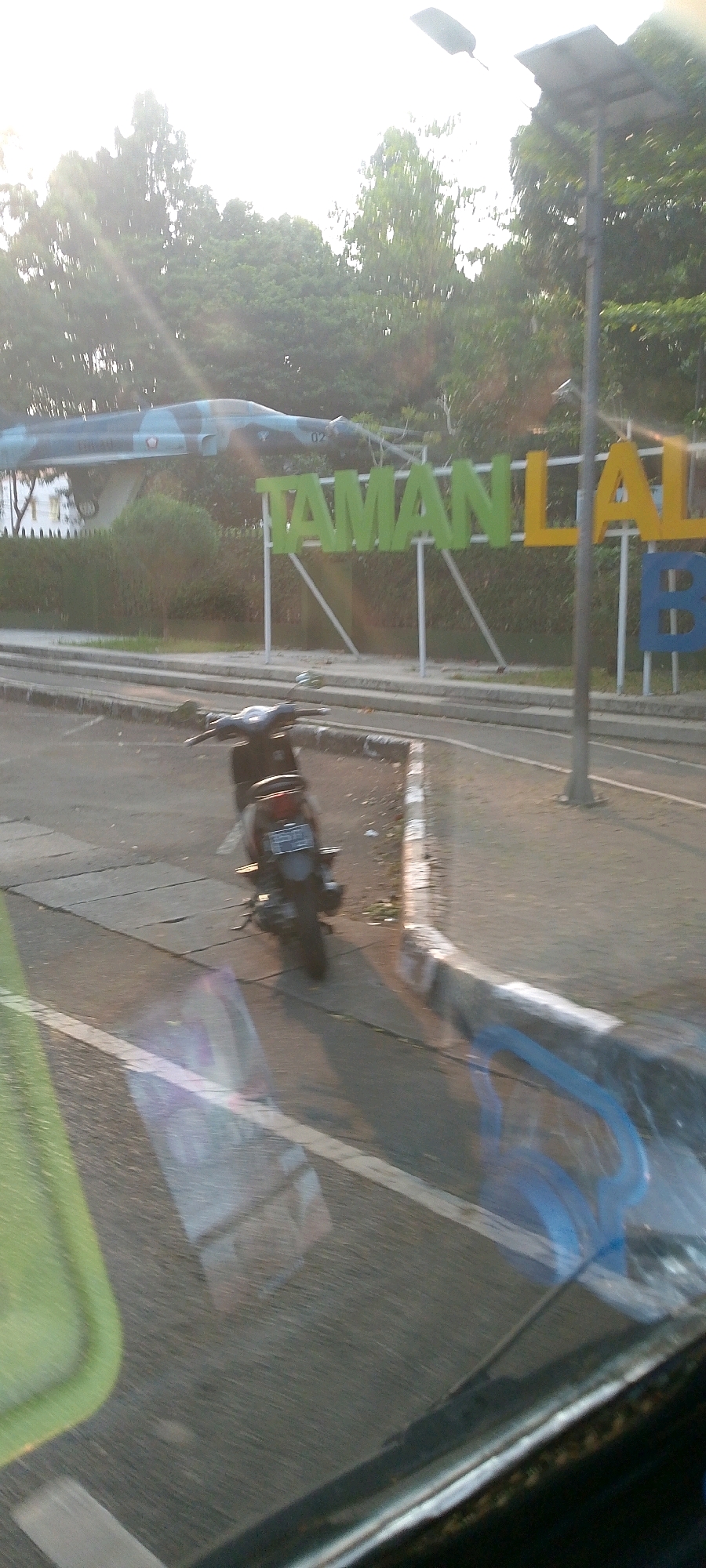 The image captures a scene in Bandung City, Indonesia, likely at a park or public space. In the background, a de-commissioned jet fighter is mounted for display, positioned above a large sign with colorful lettering that reads "TAMAN LAL". The letters are a mix of green, yellow, and blue. To the right of the sign, a modern lamppost with a solar panel is visible. The foreground features a dark-colored scooter parked on a paved area, with a license plate that is partially obscured. The environment is outdoors, with trees visible behind the jet and the sign. The lighting suggests it is either early morning or late afternoon, with sunlight casting rays through the trees. The perspective appears to be from inside a vehicle, as indicated by reflections on the surface in the lower part of the image.