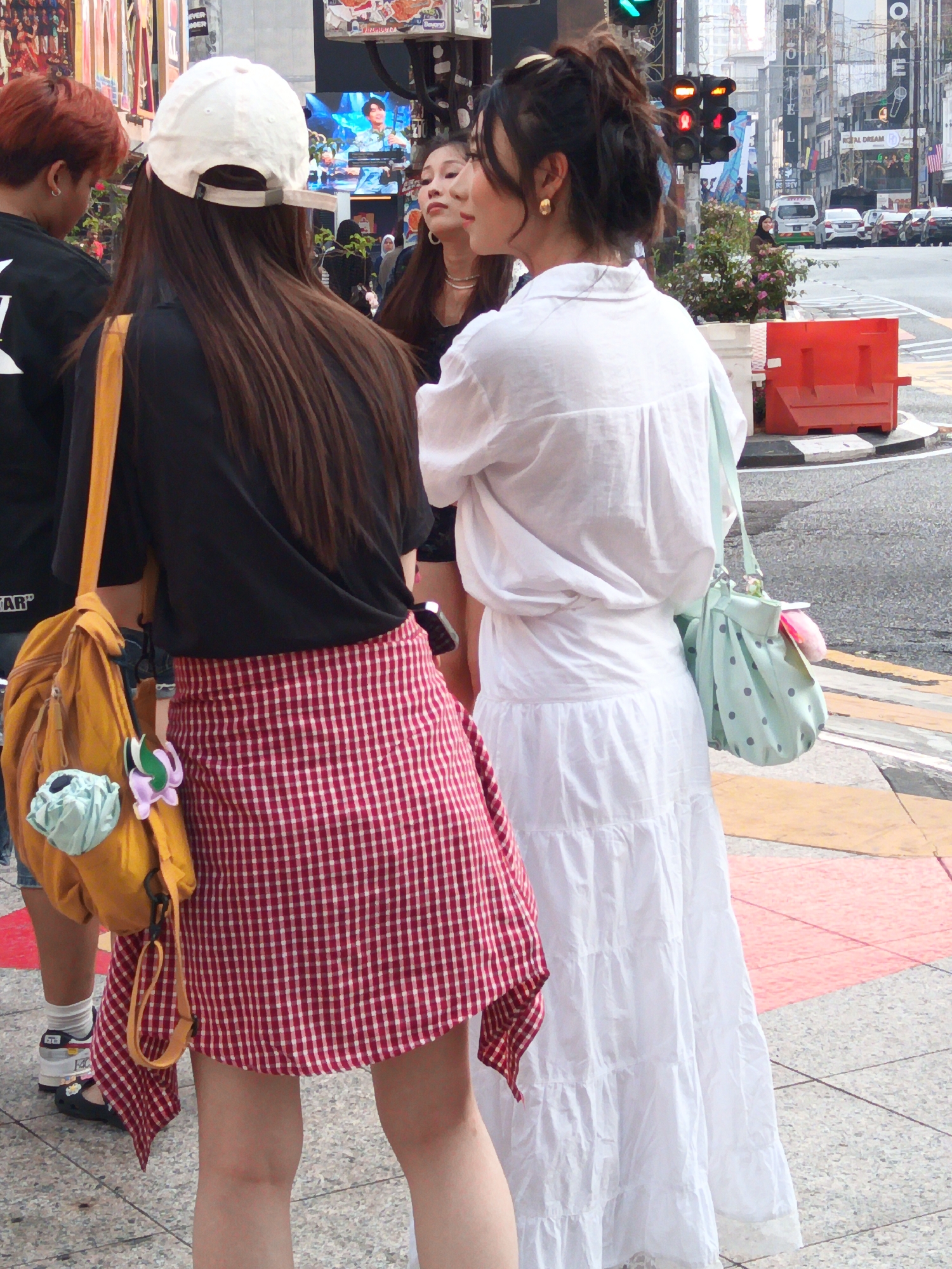 The image shows a street scene in Kuala Lumpur, Malaysia, possibly on Jalan Bukit Bintang. Two women are the primary subjects, viewed from behind and side. The woman on the left wears a black t-shirt, a red and white checkered skirt, a white baseball cap, and carries a yellow backpack adorned with two small charms. The woman on the right is wearing a white long-sleeved button-down shirt and a long tiered white skirt. She has a light blue polka-dot handbag with a light green strap. They appear to be walking or standing on a city sidewalk near a crosswalk. In the background, there are buildings, traffic lights, and vehicles, suggesting an urban environment.