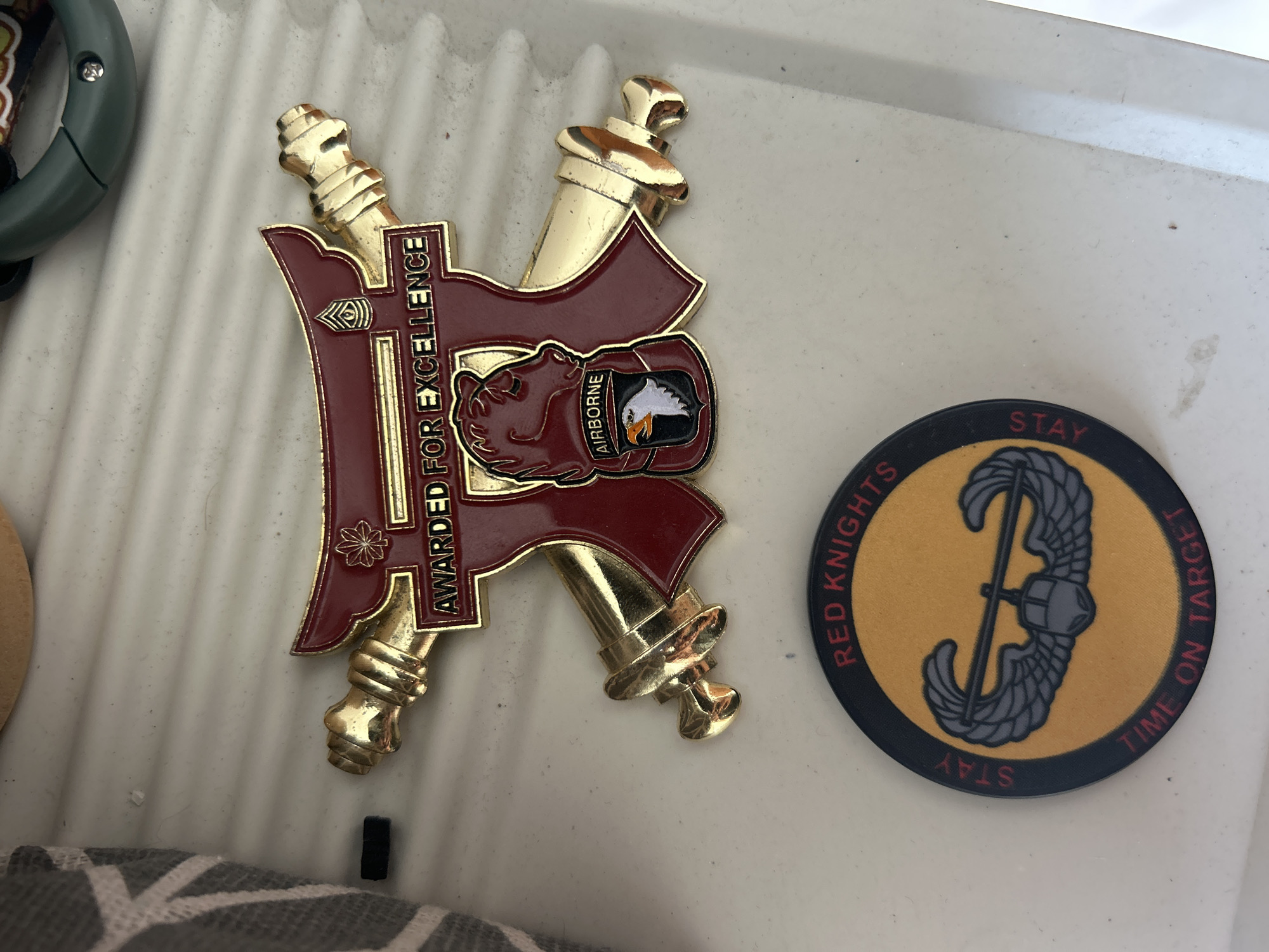The image displays two military-themed items on a white, textured surface. The larger item is a red and gold award, possibly a challenge coin or medal, featuring a stylized depiction of a bear's head, an eagle with the word "AIRBORNE" below it, and the text "AWARDED FOR EXCELLENCE." It is adorned with gold accents that resemble crossed sabers or rifles. The smaller item is a round patch or coaster with a yellow center and a black border. The yellow center contains a gray image of a parachute with a pilot's helmet in the center. The border has red text that reads "RED KNIGHTS" at the top and "STAY TIME ON TARGET" around the bottom. The items are placed on a white, slightly rippled surface, possibly a table or shelf. The background shows a hint of a gray and white striped fabric on the left and a green object in the upper right corner, which appears to be a keychain or part of a larger item. There is no indication of the city of Sukawati, Indonesia in the image.