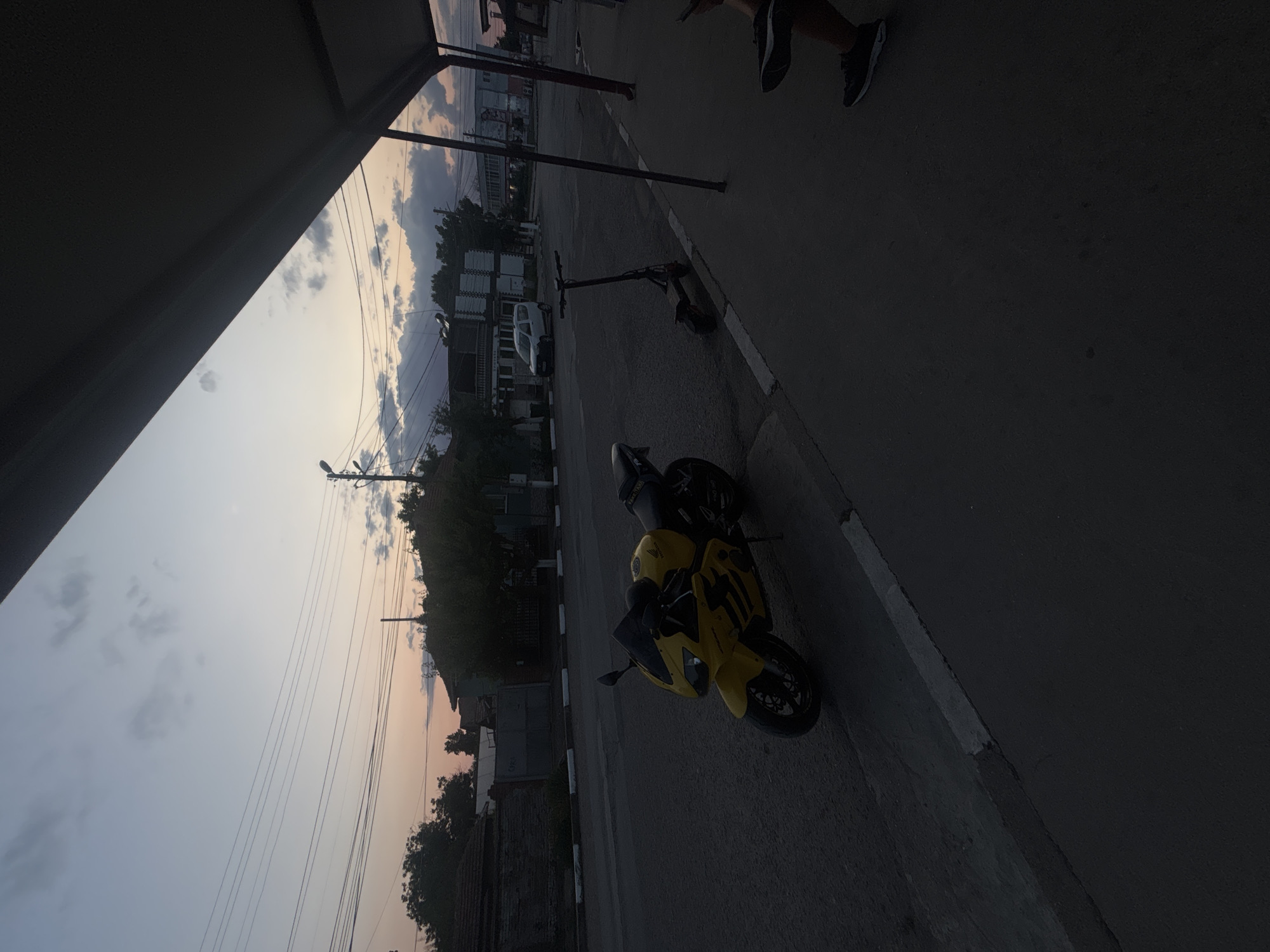 The image depicts an outdoor scene with a street in Marten, Bulgaria. The time of day appears to be dusk, as indicated by the soft, muted colors in the sky with hints of pink and orange, and the overall dim lighting. There are clouds visible, suggesting a potentially mixed weather condition, though not overtly stormy.
The primary subjects are two motorcycles parked on the asphalt road. The one in the foreground is a bright yellow sportbike, facing towards the right side of the frame. It has a sleek design with visible front forks, handlebars, and a wheel. Behind it, another motorcycle, predominantly black, is also parked.
The setting includes a street with a white and black striped curb on the left. Beyond the curb, there are buildings and trees. Some of the buildings appear to be residential, with tiled roofs. Electrical wires are strung across the sky, typical of many urban and semi-urban environments. In the background, a white van is visible, and further back, more structures, some with visible windows and balconies, are present. There's also a glimpse of what looks like a lamppost.
On the upper right side of the image, the lower legs and feet of two people wearing dark shoes are visible, suggesting they are standing or walking near the motorcycles. One person appears to be wearing dark pants and sneakers, and the other, dark pants and possibly athletic shoes. Their presence implies some form of activity is occurring, perhaps related to the motorcycles or simply a moment of rest.
The overall mood of the image is quiet and still, possibly capturing a moment of transition at the end of the day. There are no discernible emotions strongly conveyed, beyond a sense of everyday life. No visible text is present in the image.