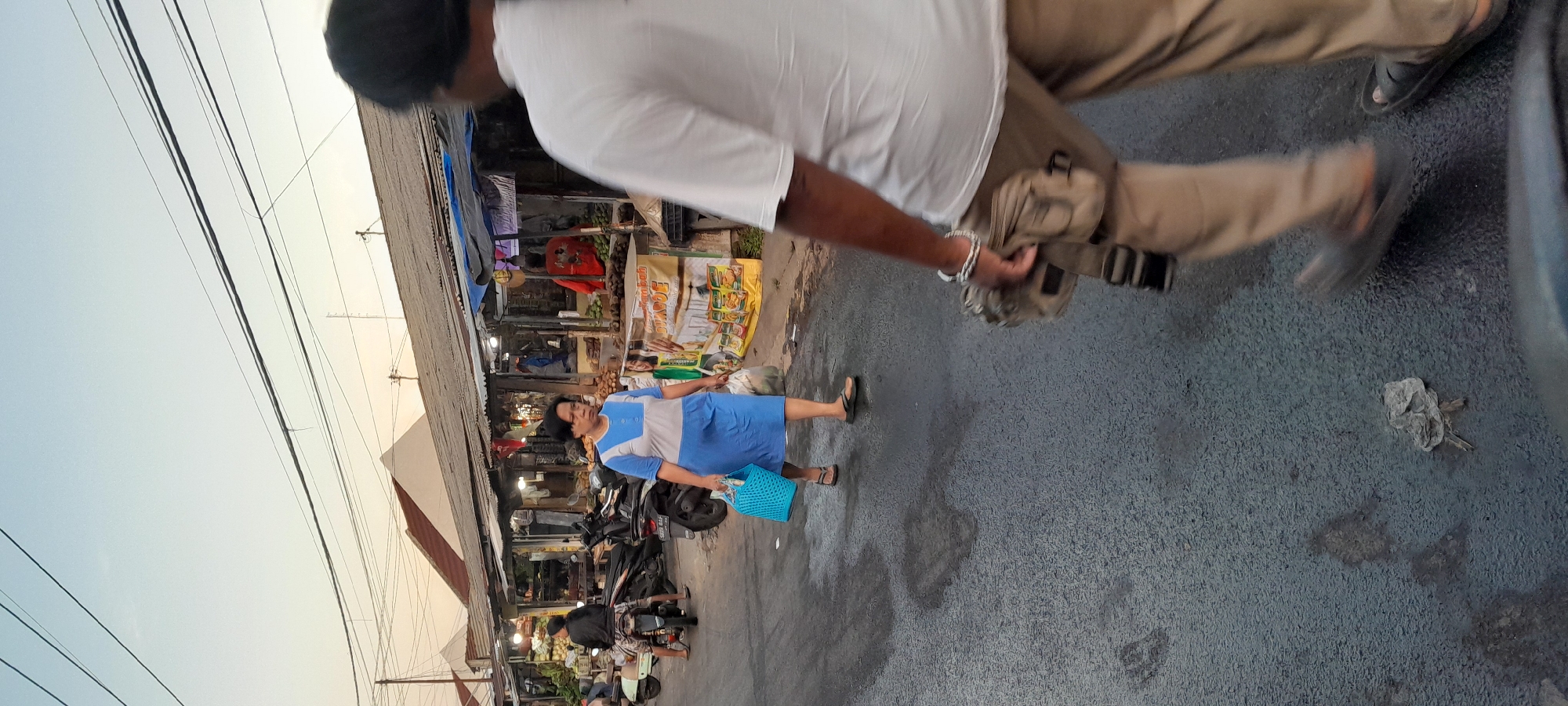 The image depicts a street scene in Rogojampi, Indonesia, likely a market area. A woman in a blue and gray dress is walking on the left side of the asphalt road, carrying a blue woven bag and a plastic bag. She is wearing sandals. In the background, stalls line the street, some with awnings. One stall displays produce, and a person in a red shirt is visible behind the counter. Motorcycles are parked along the side of the road. The sky is overcast with a pale gradient. A person, partially visible and out of focus in the foreground on the right, is walking away from the camera, wearing a white t-shirt, tan pants, and a fanny pack. Their left foot, wearing a dark sandal, is in motion. The road surface shows some wet patches and debris.