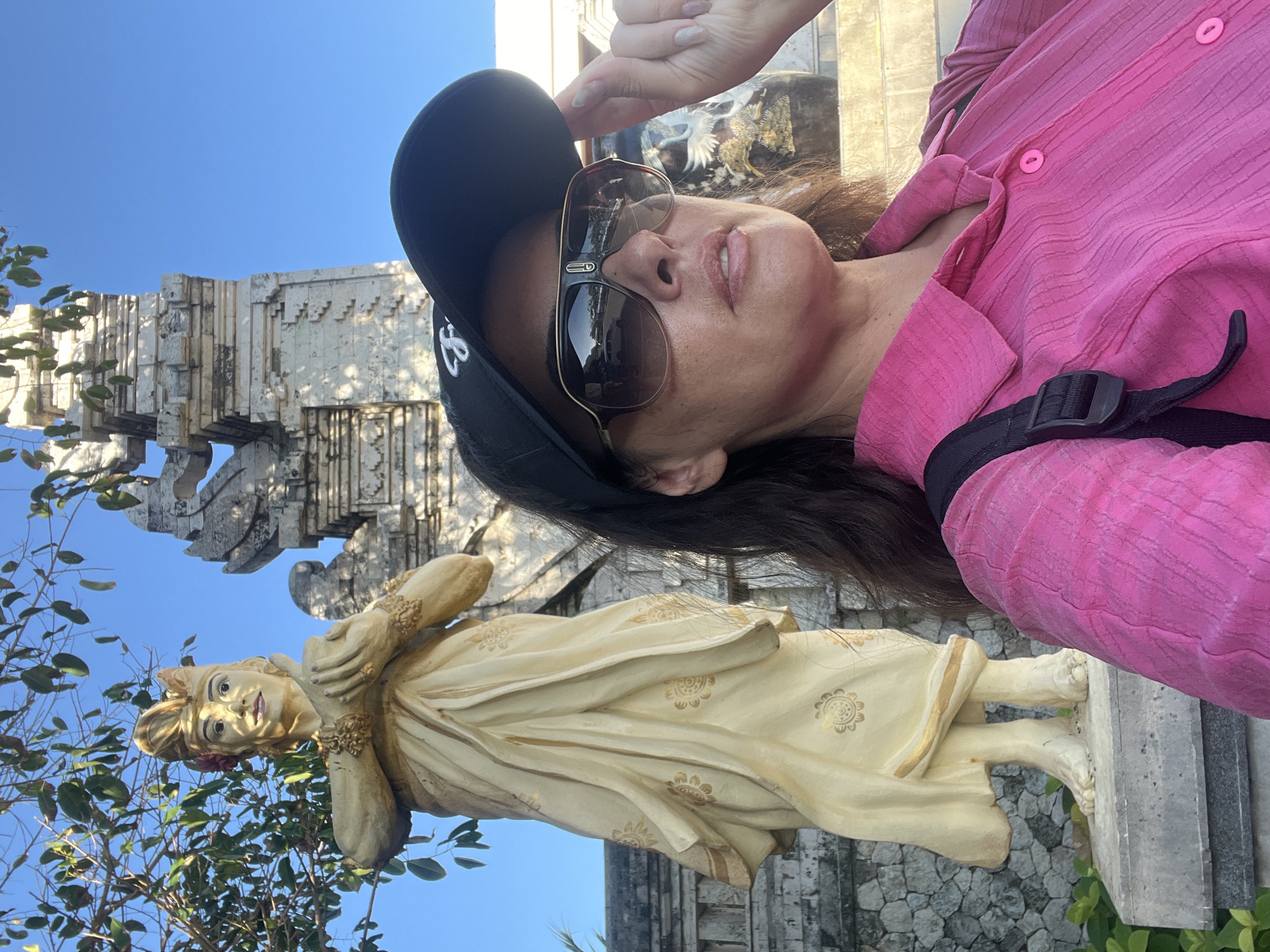 The media file depicts a woman taking a selfie in an outdoor setting, with a statue and architectural elements in the background. The woman is wearing a pink shirt, sunglasses, and a black baseball cap. Her right hand is raised, touching the brim of her cap. She has long, dark hair and appears to be looking at the camera.
In the background, there is a light-colored statue of a person, possibly a deity or historical figure, adorned with gold accents. The statue is dressed in flowing robes with intricate patterns. Behind the statue, a stone structure with detailed carvings is visible, suggesting a temple or historical monument. The scene is set against a clear blue sky, indicating a sunny day. Some greenery from trees is also present in the foreground.
The overall mood is casual and exploratory, as if the woman is on a trip or visiting a tourist attraction. The lighting suggests it is daytime, likely morning or afternoon, given the bright sunlight casting shadows. The architectural style and the statue hint at a location with rich cultural or historical significance, consistent with the provided location context of Kuta Selatan, Indonesia, which is known for its temples and cultural sites. There is no visible text in the image.