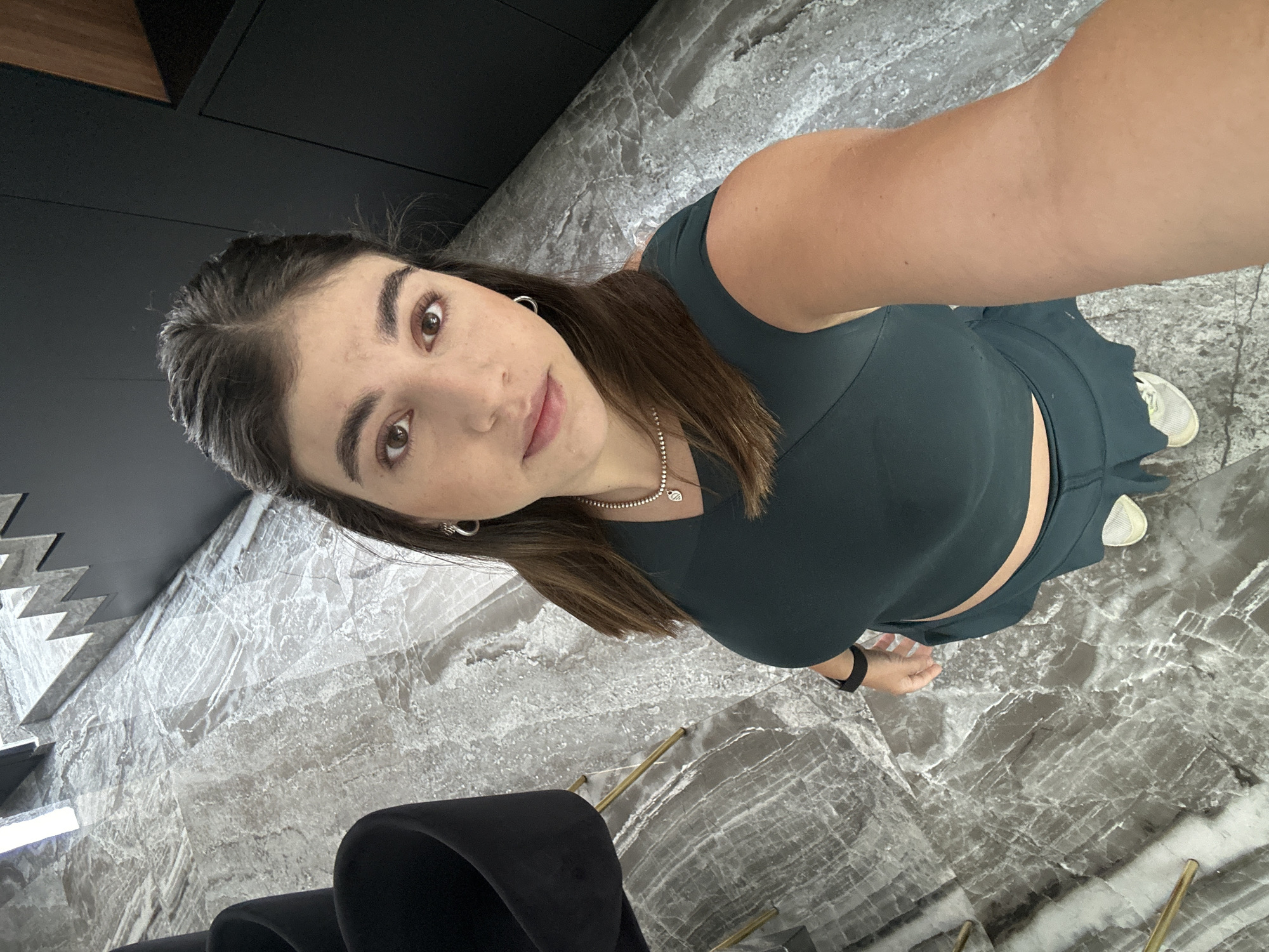 A young woman is taking a selfie. She is wearing a dark green workout set consisting of a tank top and high-waisted shorts. She has brown hair and is wearing a silver necklace and hoop earrings. She is also wearing a black smartwatch on her left wrist. She is standing indoors on a marble floor. In the background, there is a dark cabinet and chairs. The location is Puebla, Mexico.