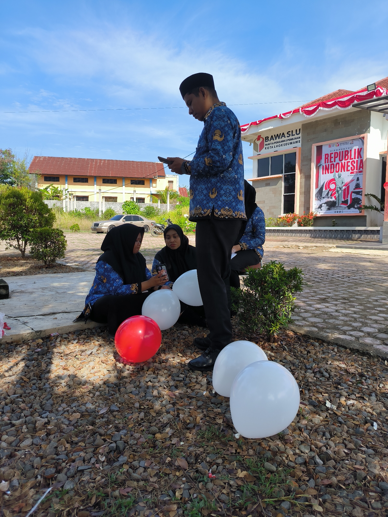 Four individuals, three women wearing black hijabs and blue patterned shirts, and one man wearing a blue patterned shirt and black peci, are present outdoors. Two women are seated on the ground, looking at their phones, while the man stands, also looking at his phone. Two white balloons and one red balloon are on the ground in front of them. Behind them is a building with "BAWASLU PANITIA PENGAWAS PEMILIHAN KOTA LHOKSEUMAWE" signage and a large poster for "REPUBLIK INDONESIA". The environment includes a paved walkway, gravel, and some green bushes, with a building and trees in the background. The sky is blue with some clouds.