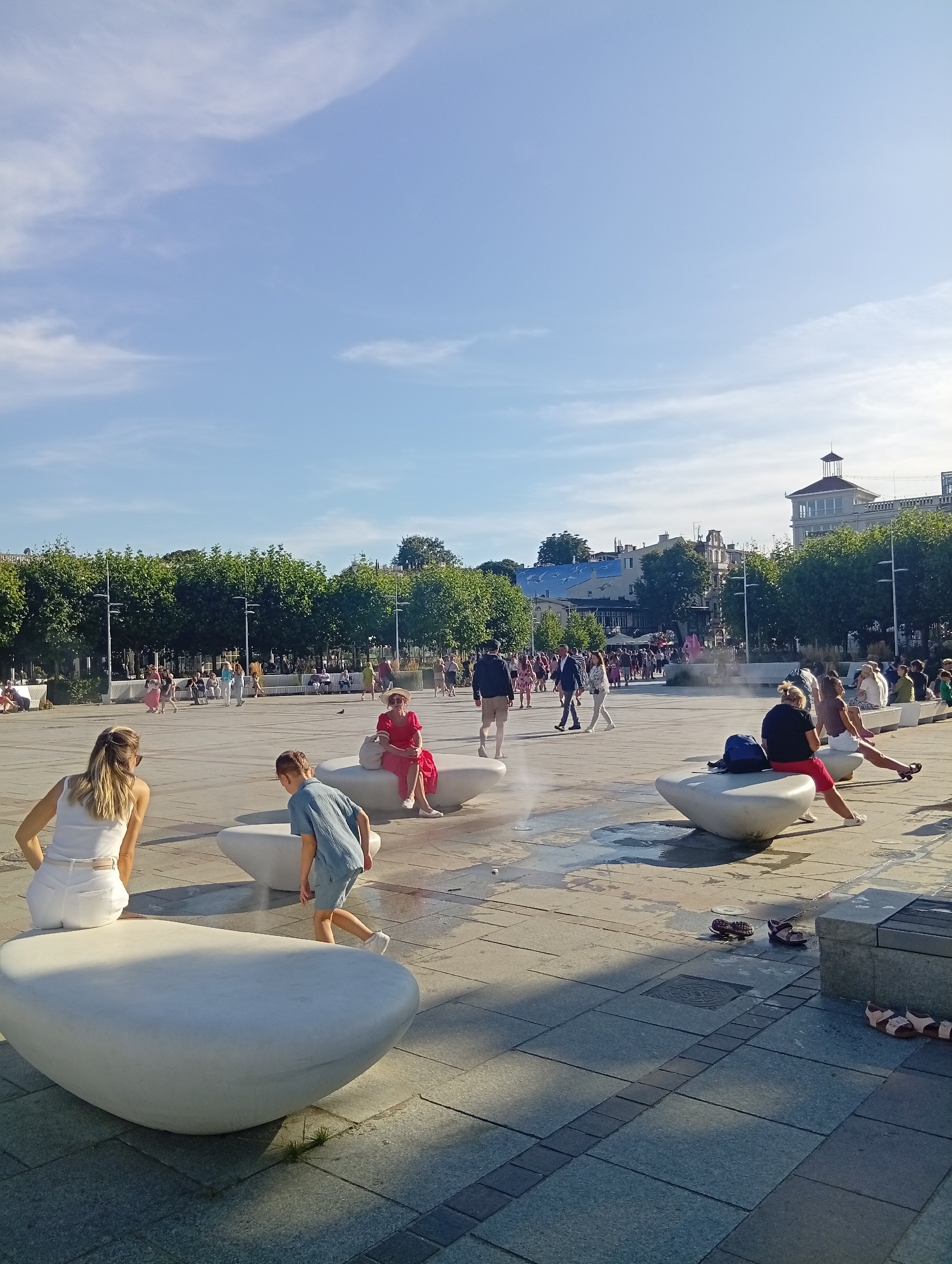 The image captures a sunny day in the city of Sopot, Poland, likely on a public square. The scene is populated with numerous people engaged in various activities. In the foreground, a woman with blonde hair is seen from behind, wearing a white top and white shorts, appearing to be seated or crouching. Beside her, a young boy in a blue t-shirt and shorts is walking away from the camera, towards some water features that create a mist.
Several large, white, egg-shaped seating structures are scattered across the paved square. One of these structures is occupied by a woman in a vibrant red dress, seated with her legs crossed, wearing a straw hat. Another seating structure to the right is occupied by two women, one in a black top and red shorts, and the other in a white top and skirt.
Further into the square, a diverse crowd of people are walking, sitting on benches, and interacting. In the background, a line of mature trees forms a green canopy, with lampposts interspersed. Behind the trees, urban buildings are visible, including a prominent building with a tower and a blue-roofed structure. Water jets are active, creating mist and spraying in some areas of the square, suggesting a public fountain or water play area. The ground is paved with large, gray square tiles, with some areas appearing wet from the water features. Several pairs of sandals and flip-flops are scattered on the pavement. The sky is blue with scattered white clouds.